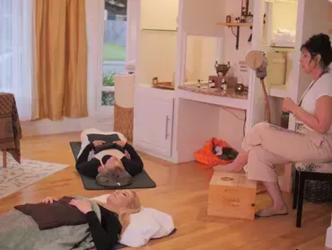 People lying on yoga mats in a bright room while an instructor sits nearby with a notepad.