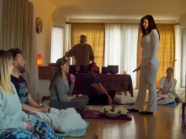 People gathered in a sunlit living room, sitting and standing around a central table.