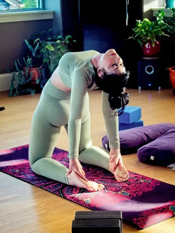 Person in a green outfit doing a deep backbend on a yoga mat in a room with cushions and plants