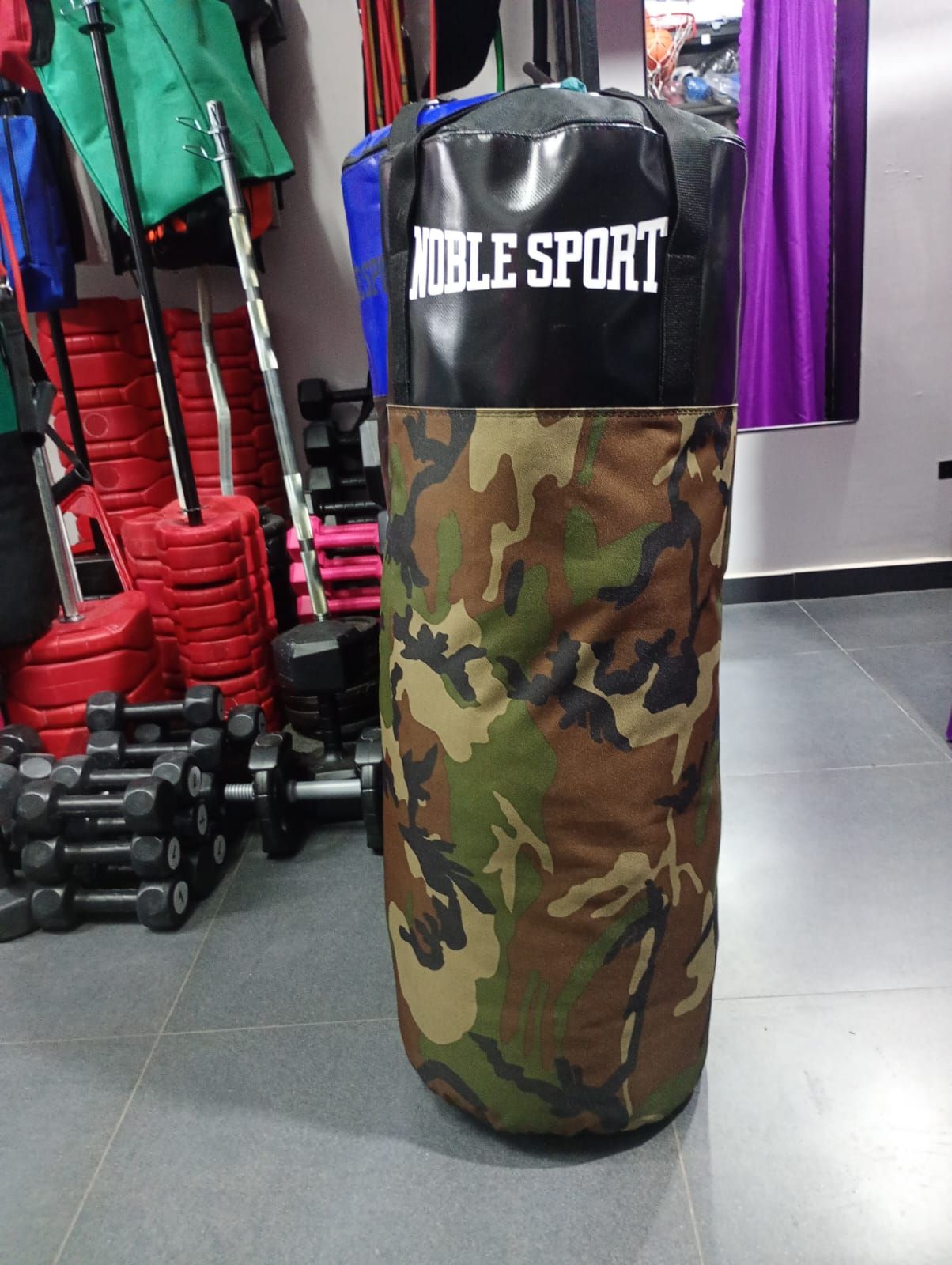 Un saco de boxeo de camuflaje de la marca Noble Sport en un gimnasio, con pesas al fondo.
