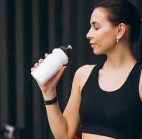 Mujer con top deportivo negro sosteniendo una botella de agua blanca, mirándola contra un fondo oscuro.