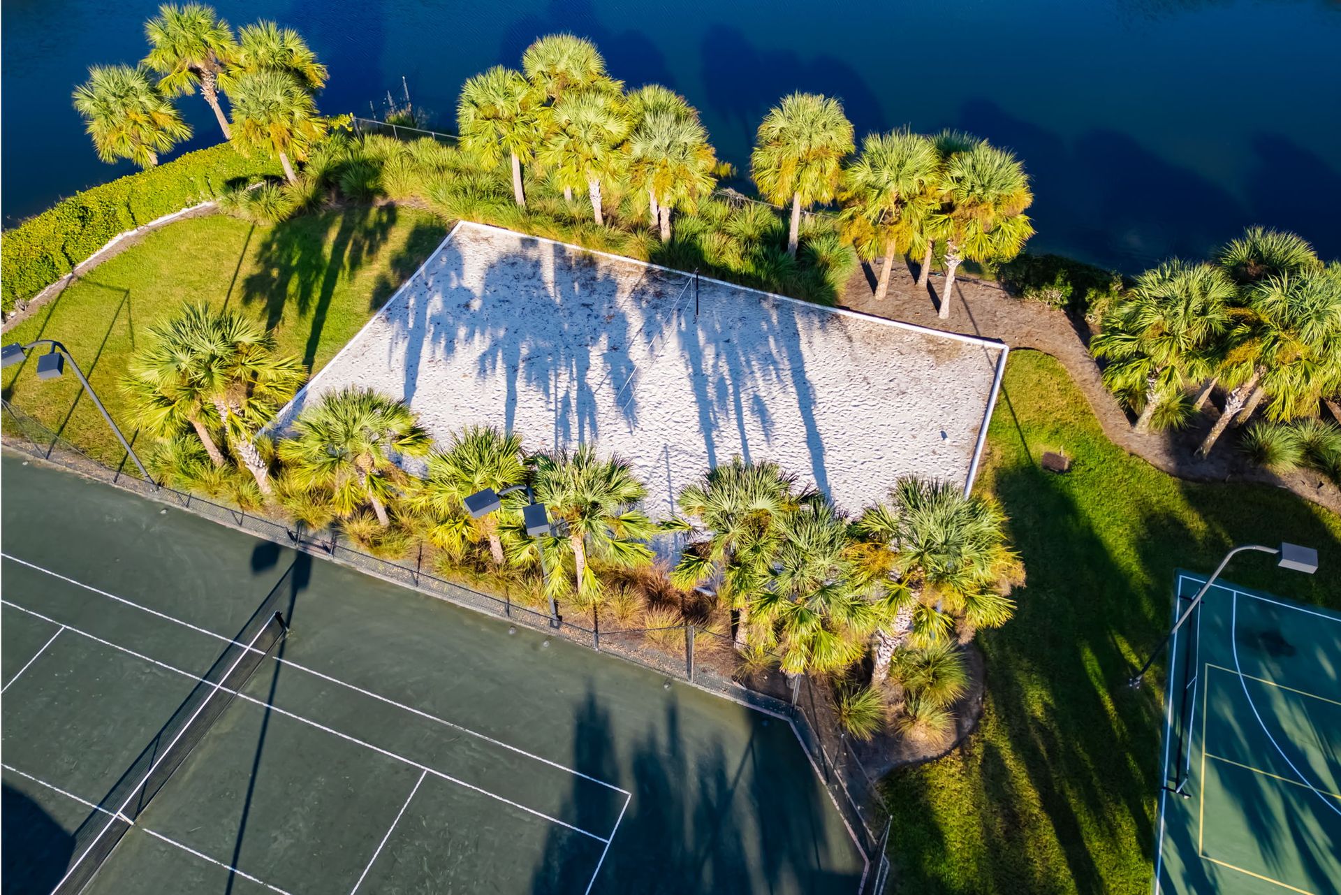 Aerial view of a tennis court beside a white-roofed building and palm trees on a sunny day