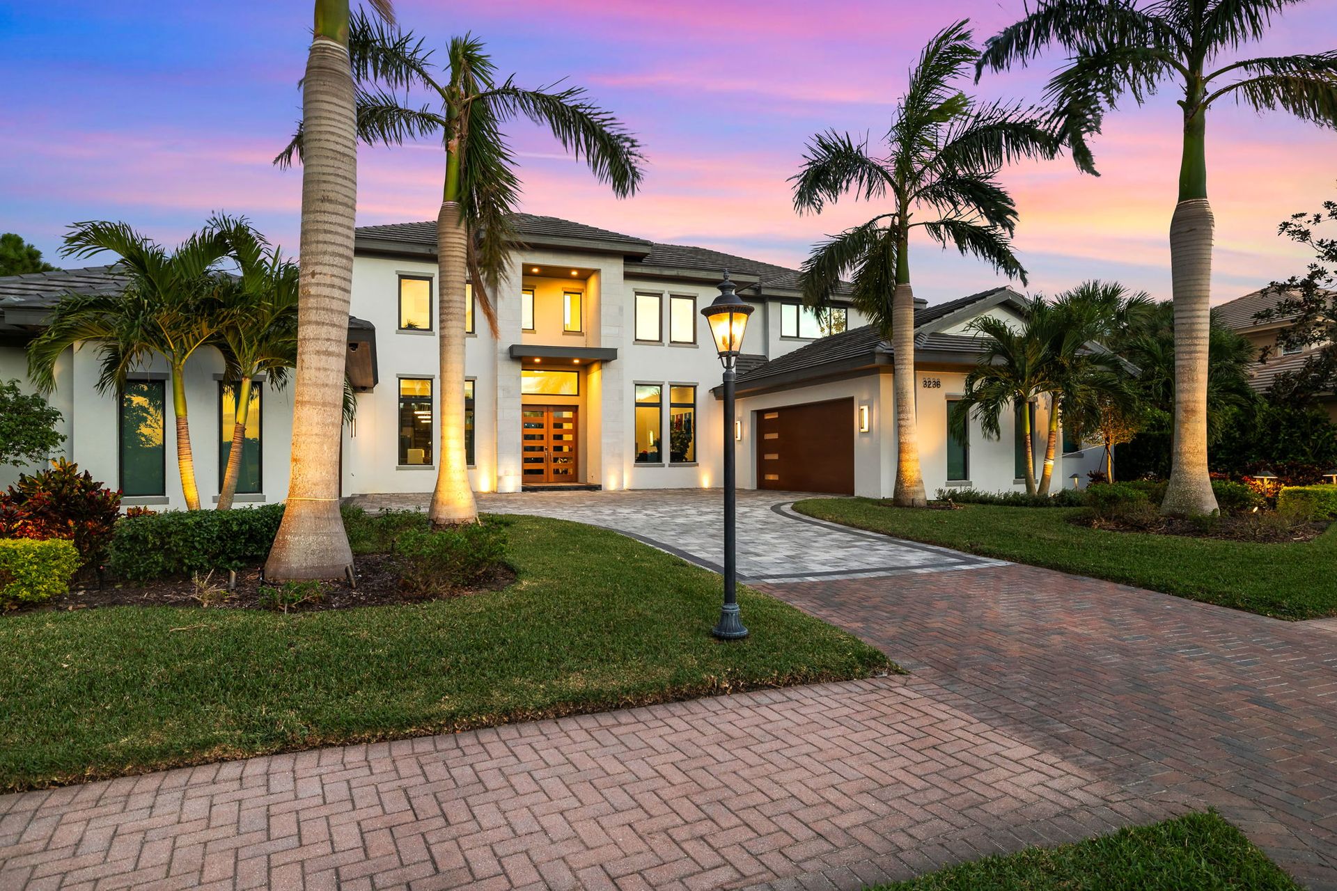 Luxury two-story house with palm trees, lit windows, and a brick driveway at sunset
