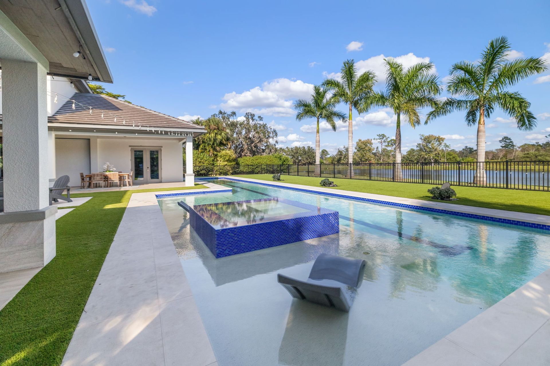 Modern backyard pool with blue tile, palm trees, and a sunny patio beside a house