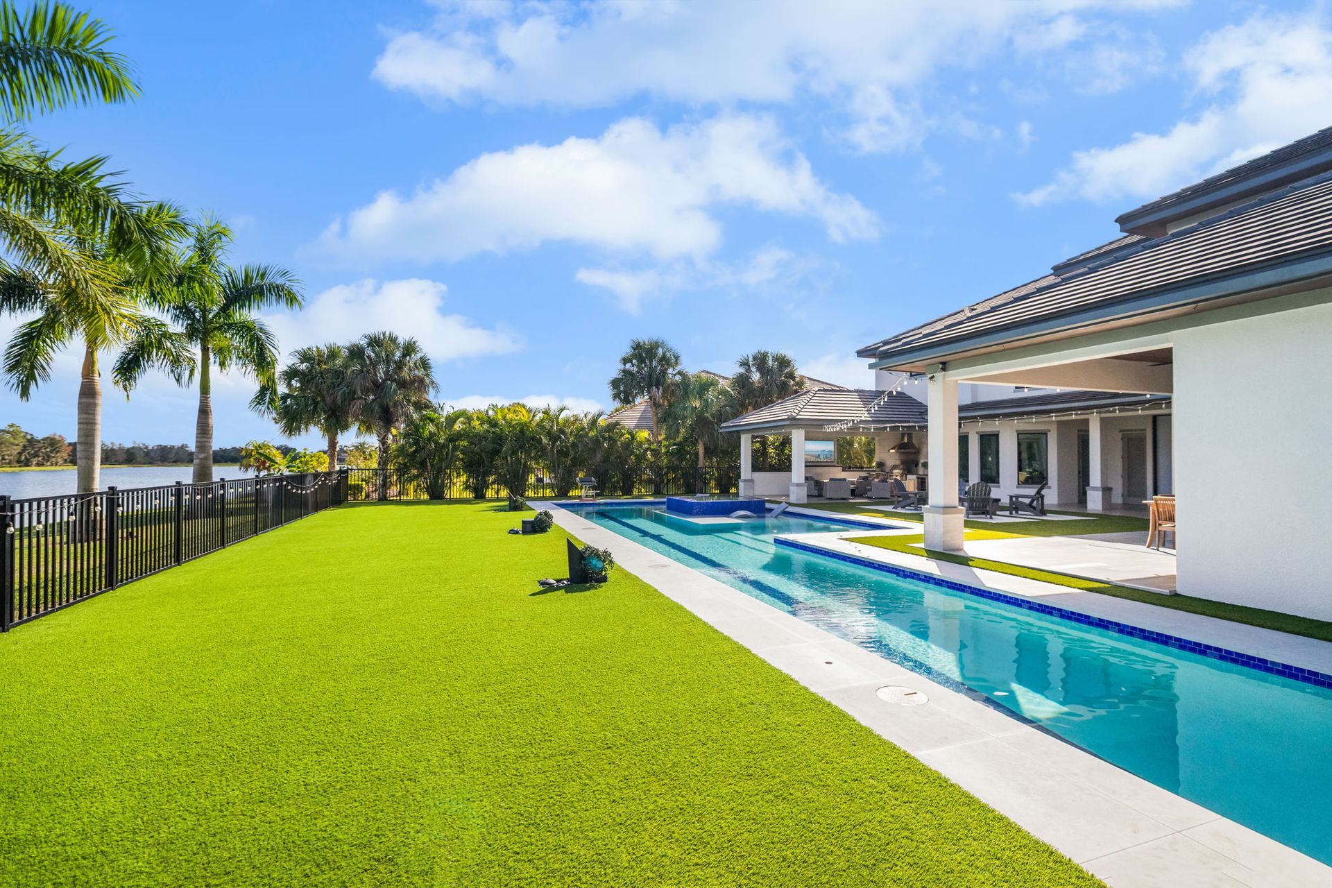 Luxury backyard with pool, lush green lawn, palm trees, and a white house under a blue sky
