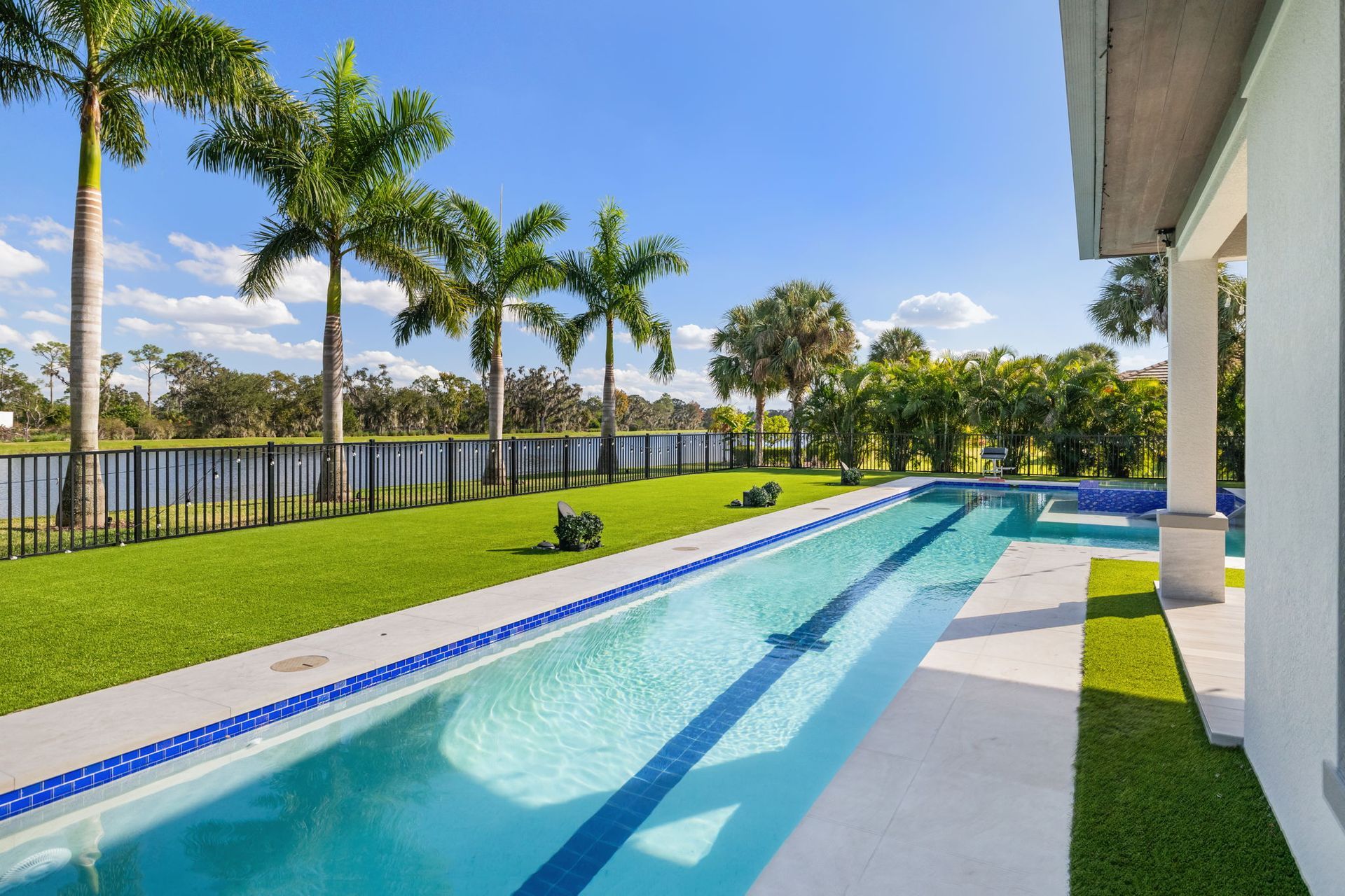 Modern backyard with a long blue pool, green lawn, palm trees, and a waterfront view