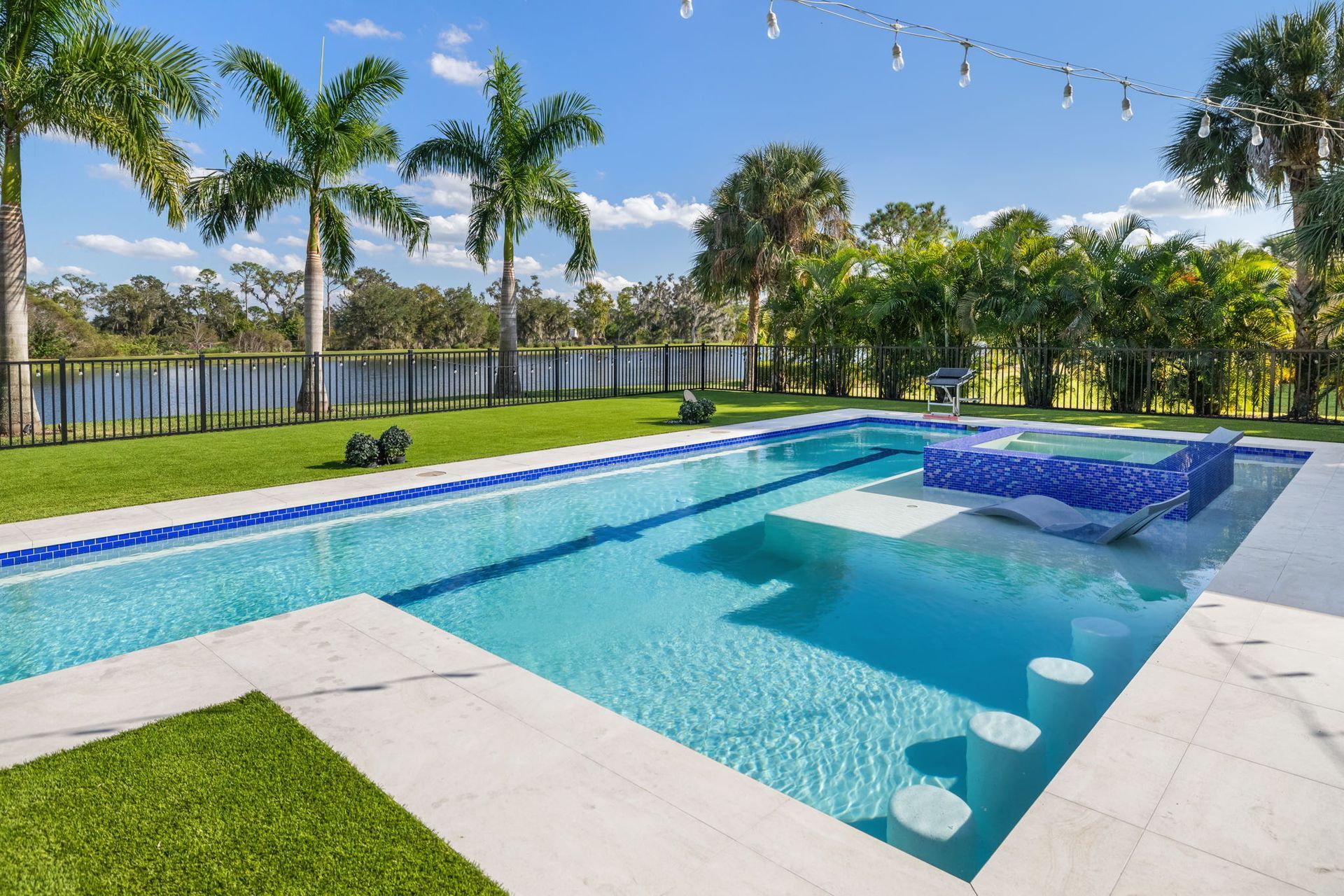 Backyard swimming pool with blue water, white deck, palm trees, and a grassy lawn by a waterfront.
