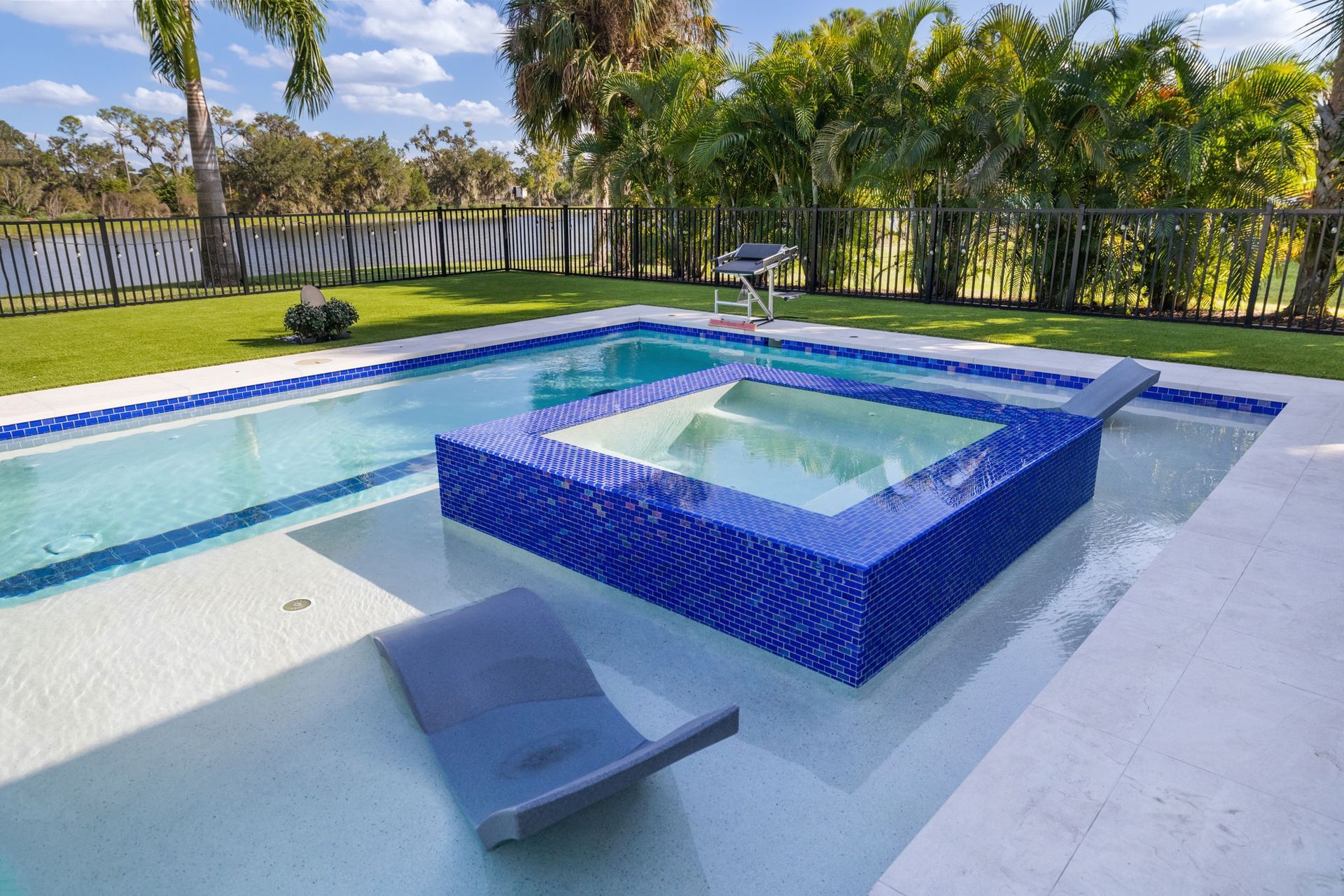 Modern backyard pool with blue-tiled square spa, shallow water, and a lounge chair under sunny skies