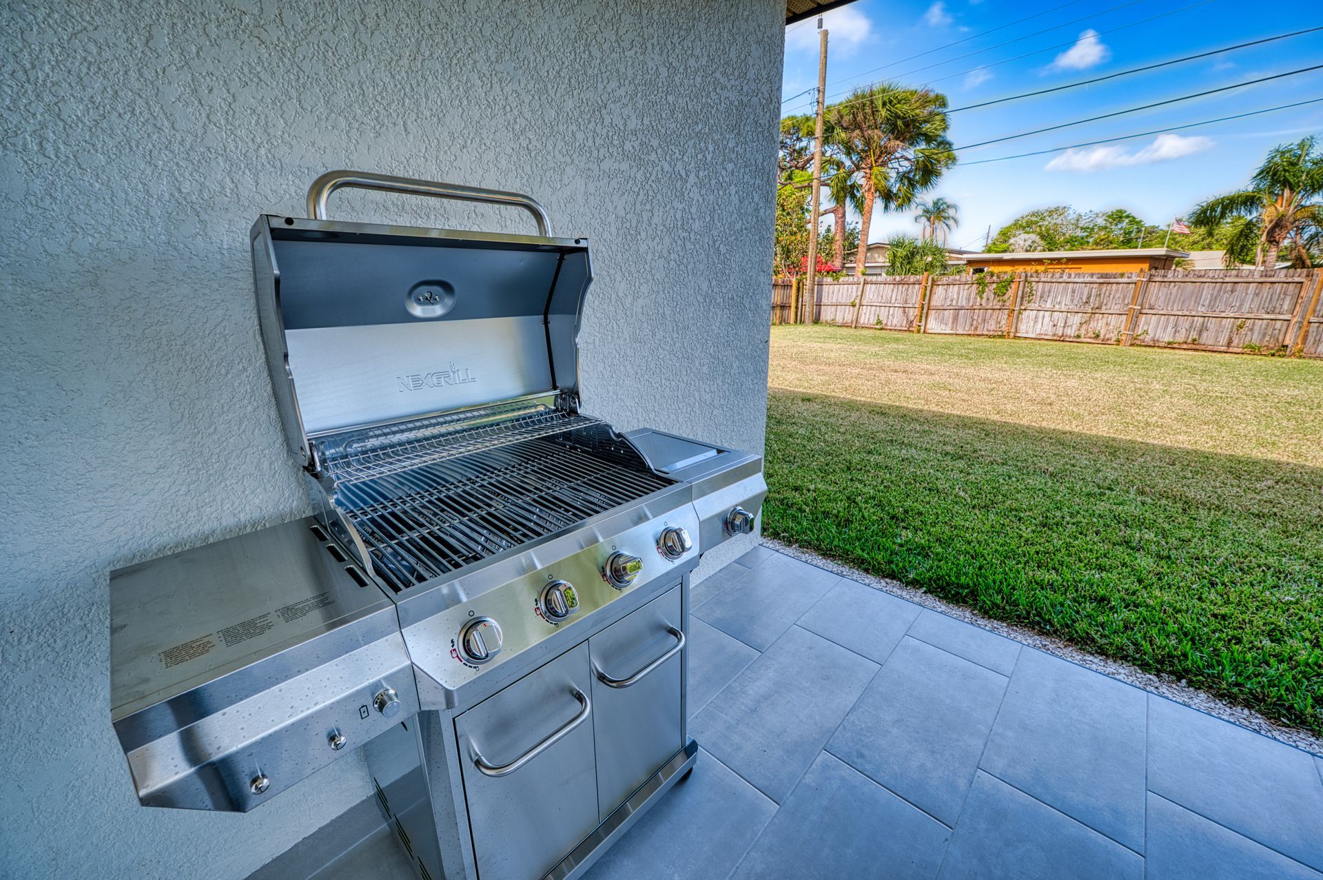 Stainless steel gas grill on a patio beside a green lawn and stone wall under a blue sky