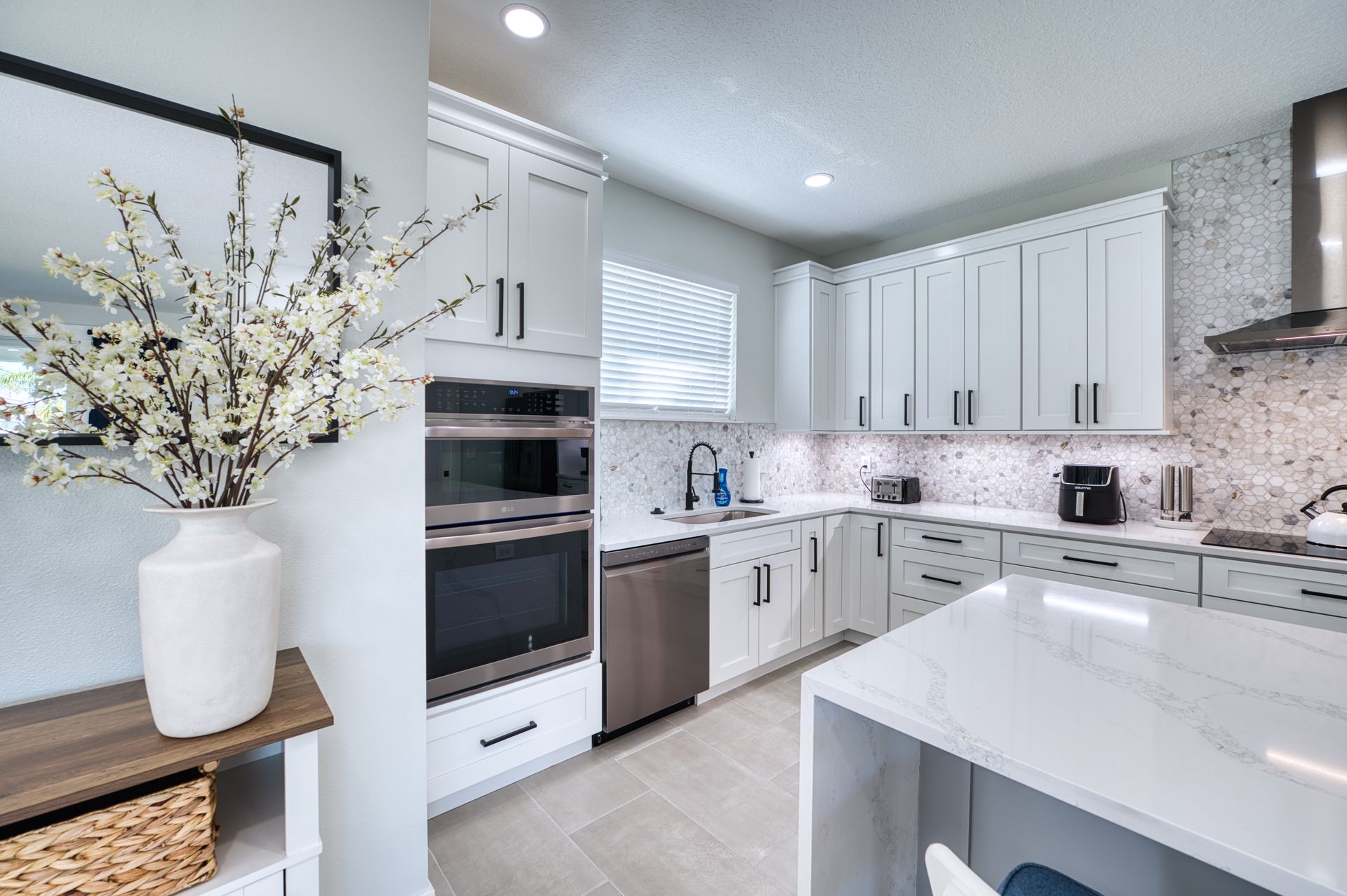 Bright modern white kitchen with marble countertops, built-in oven, and a vase of white flowers