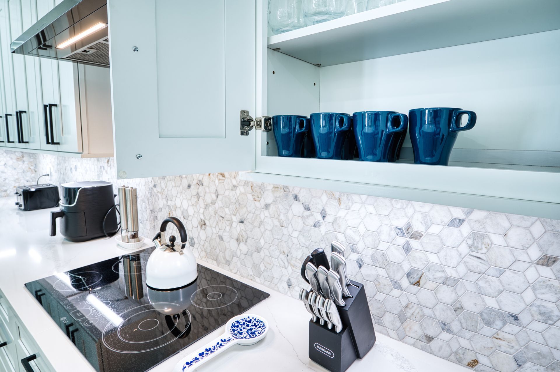Modern white kitchen with black stovetop, kettle, knife block, and blue mugs on the shelf