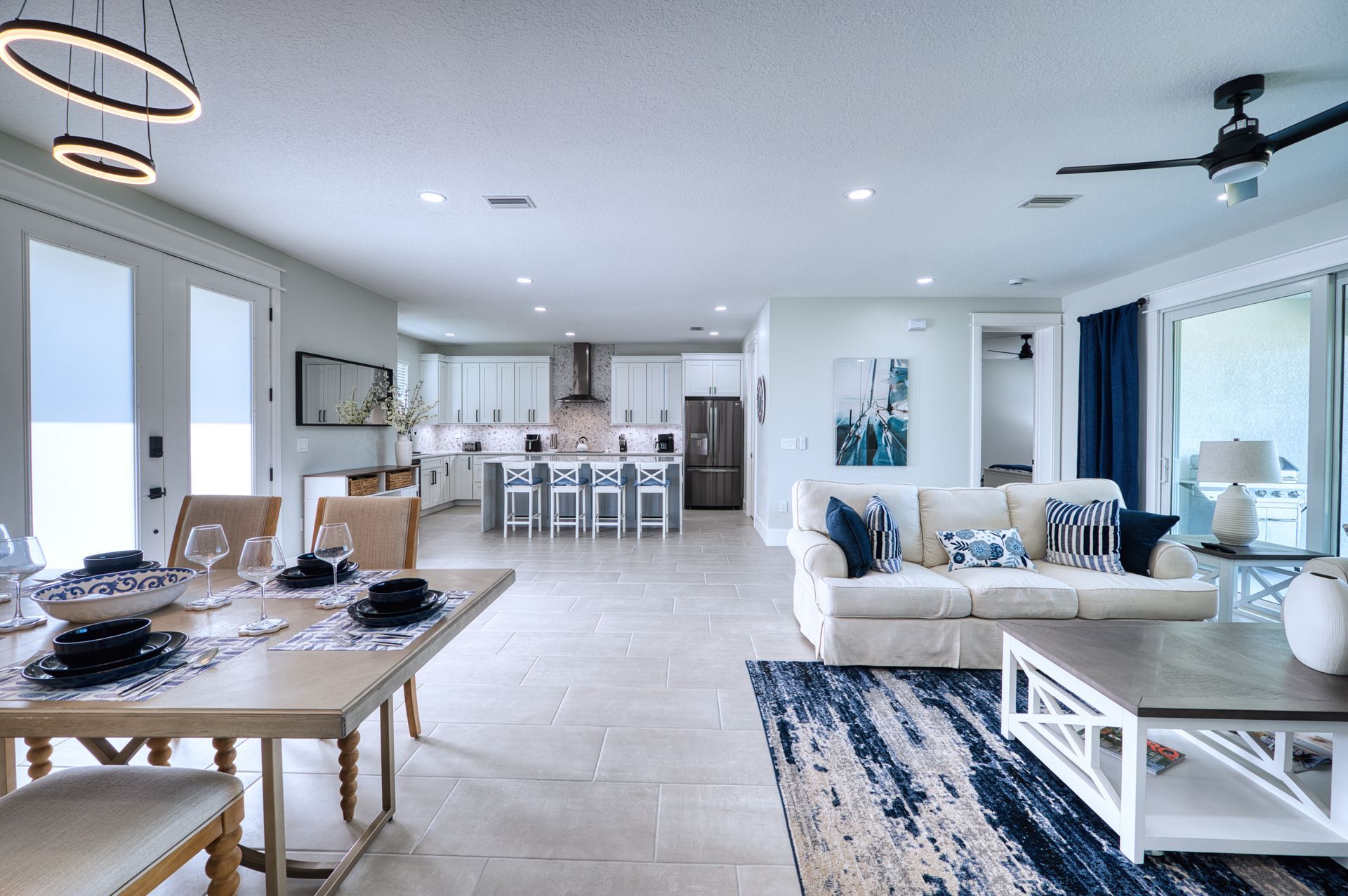 Bright open-concept living room and kitchen with white decor, blue accents, and a dining table by large windows