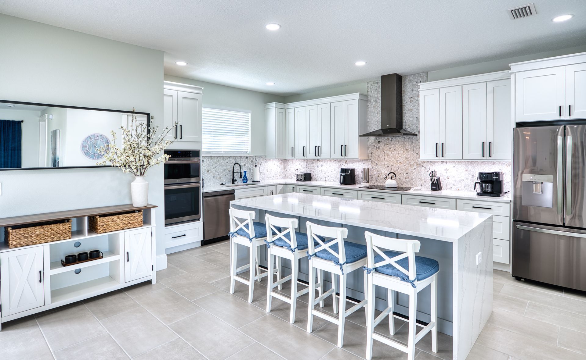 Bright modern white kitchen with island, blue stools, stainless steel appliances, and tiled floor
