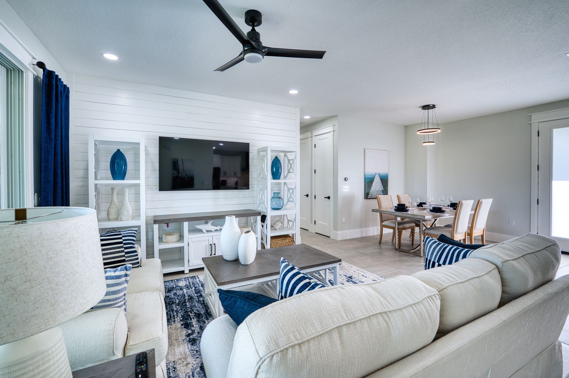 Bright modern living room with cream sofa, wall TV, blue accents, and dining area beyond.