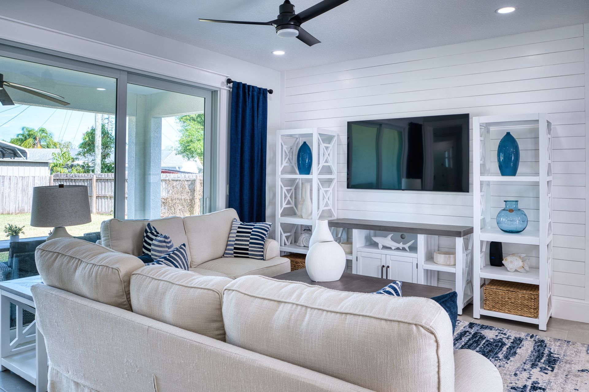 Bright living room with beige sofa, wall-mounted TV, white shelving, and blue accents near large sliding doors