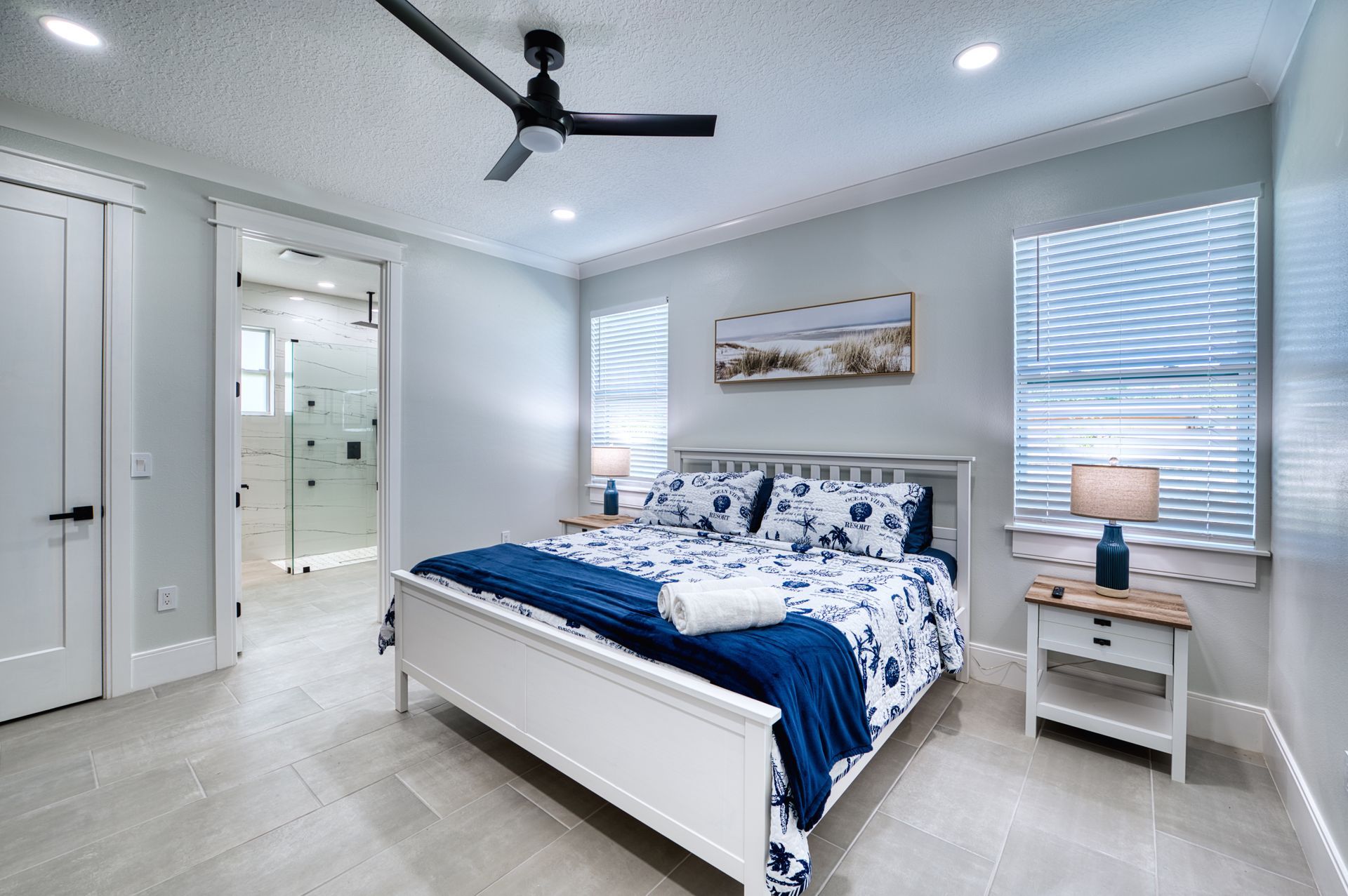 Bright bedroom with white bed, blue bedding, ceiling fan, and window blinds