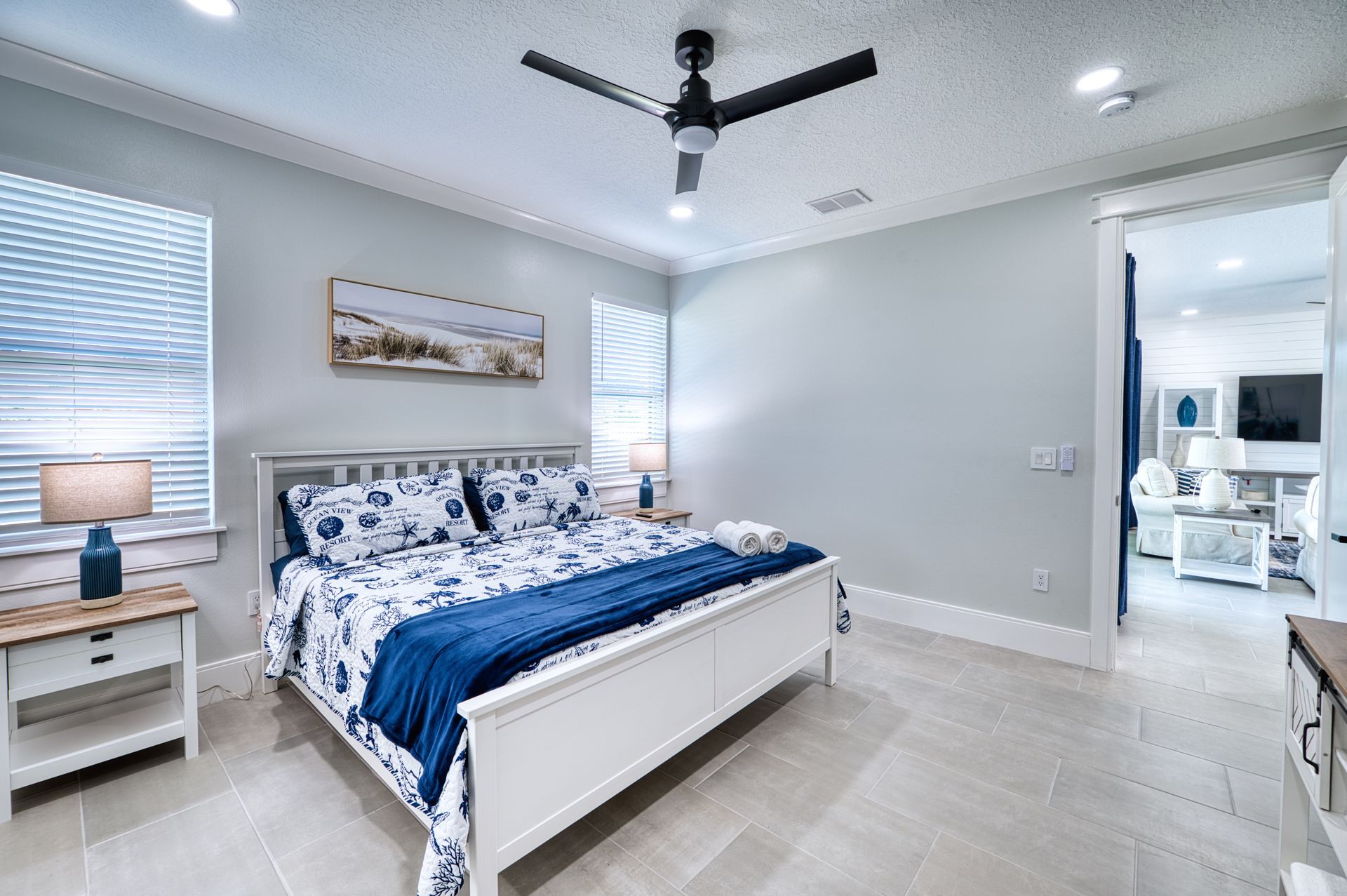 Bright bedroom with white bed, blue bedding, ceiling fan, and windows with blinds.