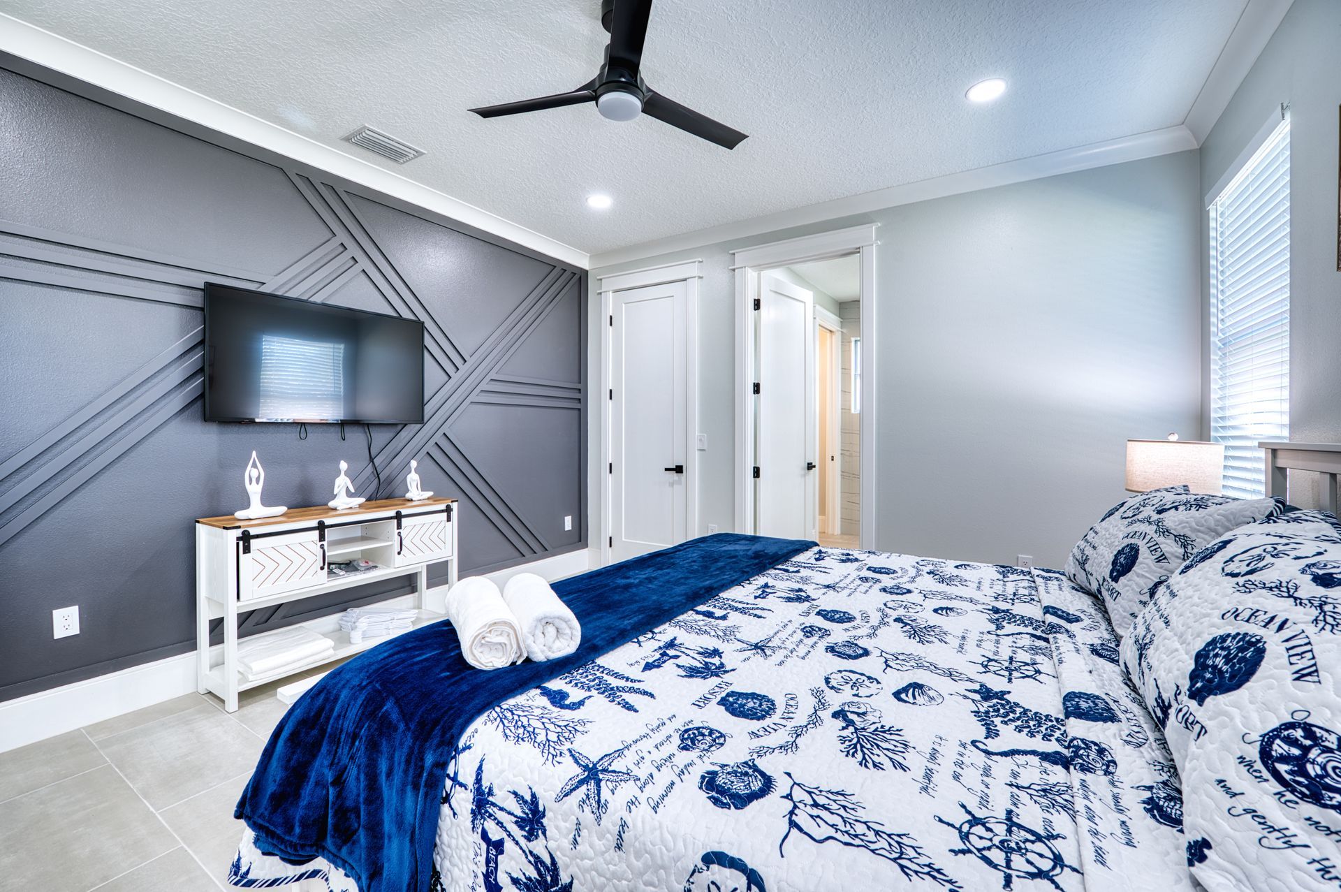 Bright bedroom with blue-and-white bedding, wall-mounted TV, ceiling fan, and closet doors.