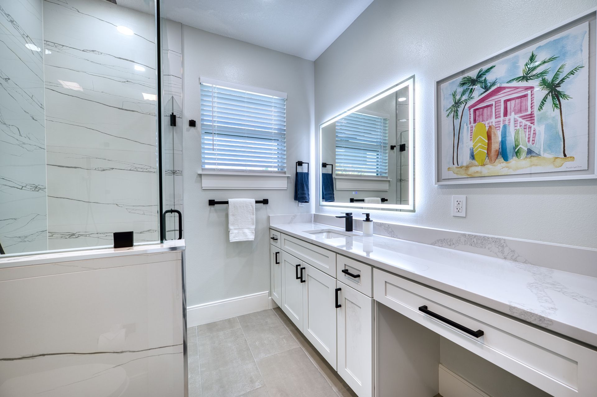 Modern bathroom with white vanity, glass shower, lit mirror, and framed artwork on the wall