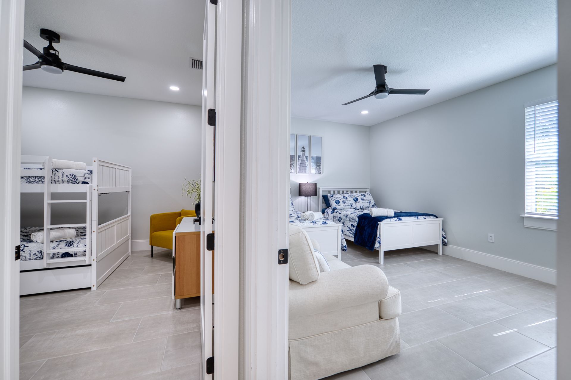 Bright bedroom with bunk beds, a white chair, and a ceiling fan; another room with a blue bed is visible.