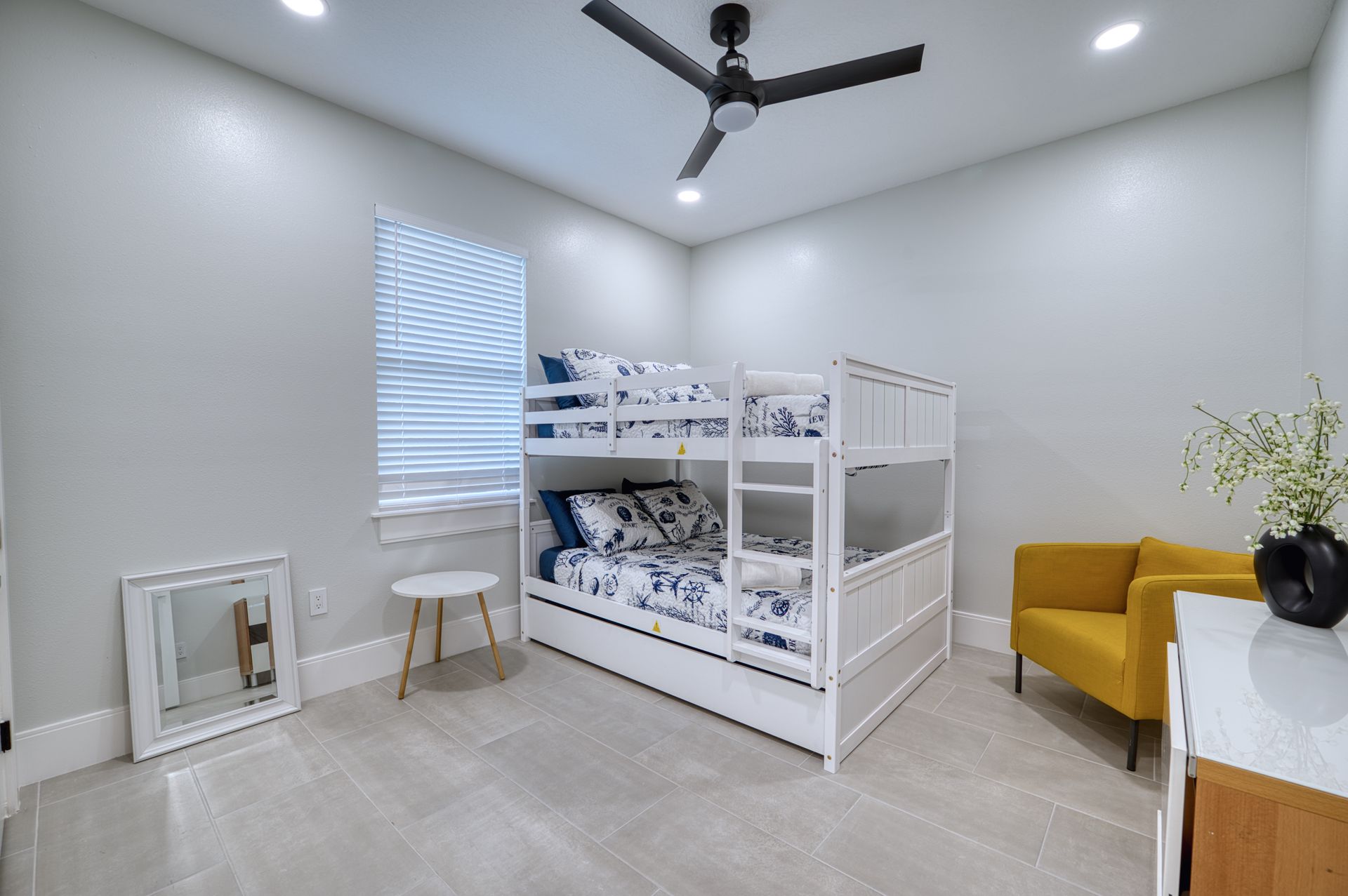 Bright shared bedroom with white bunk bed, mustard chair, and ceiling fan in a minimalist room