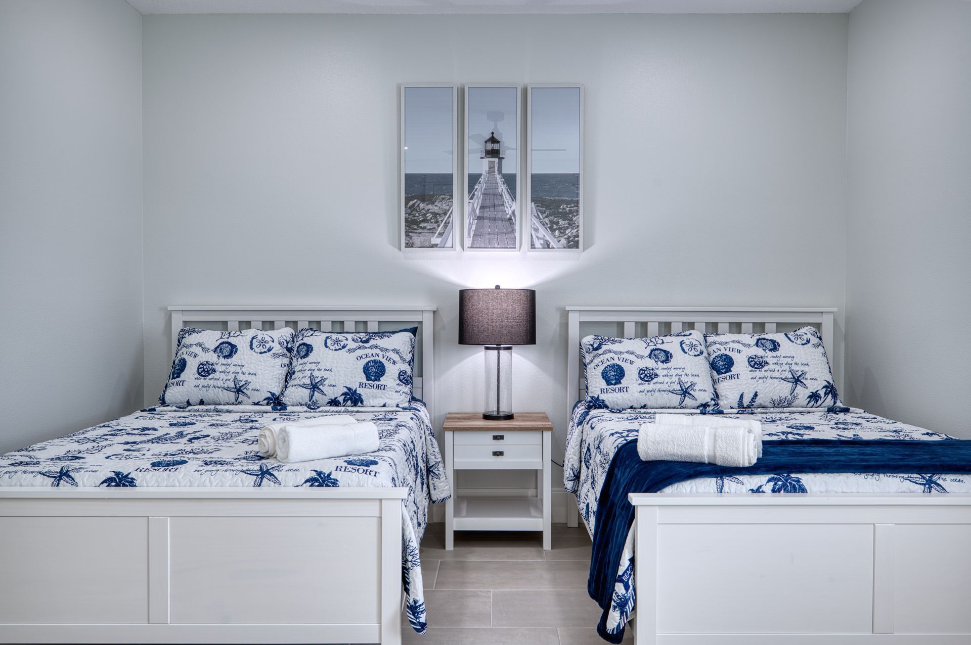 Two white twin beds with blue floral bedding in a bright bedroom, with a lamp and wall art between them
