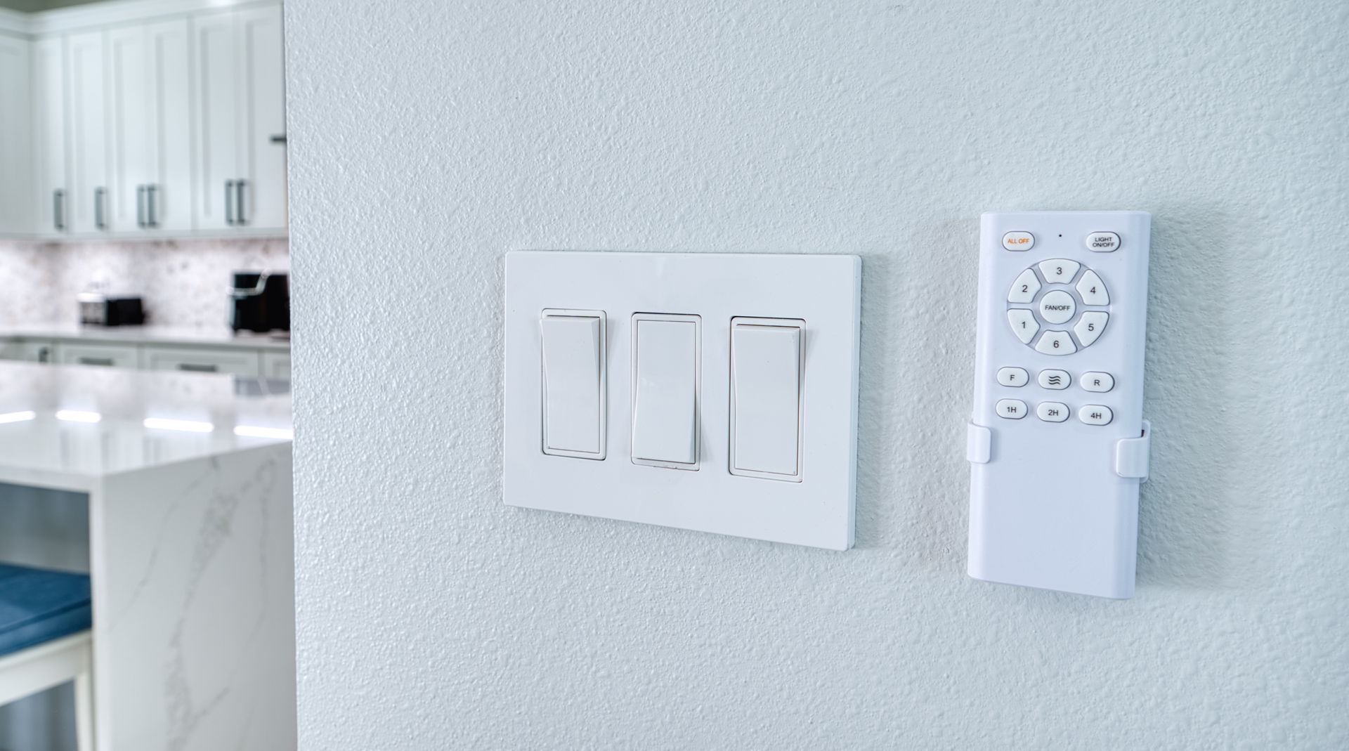 White light switches and a wall-mounted remote on a textured wall near a bright kitchen.