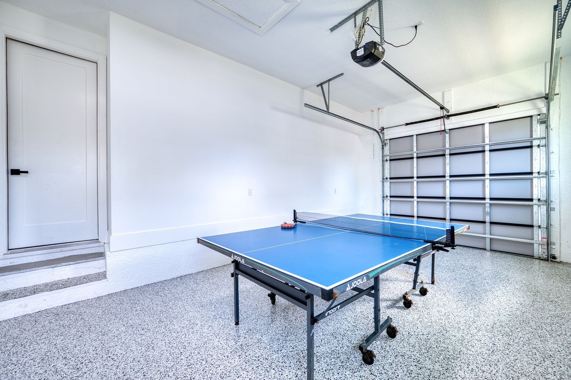 Spacious garage with a blue ping-pong table, speckled floor, and open storage shelves.