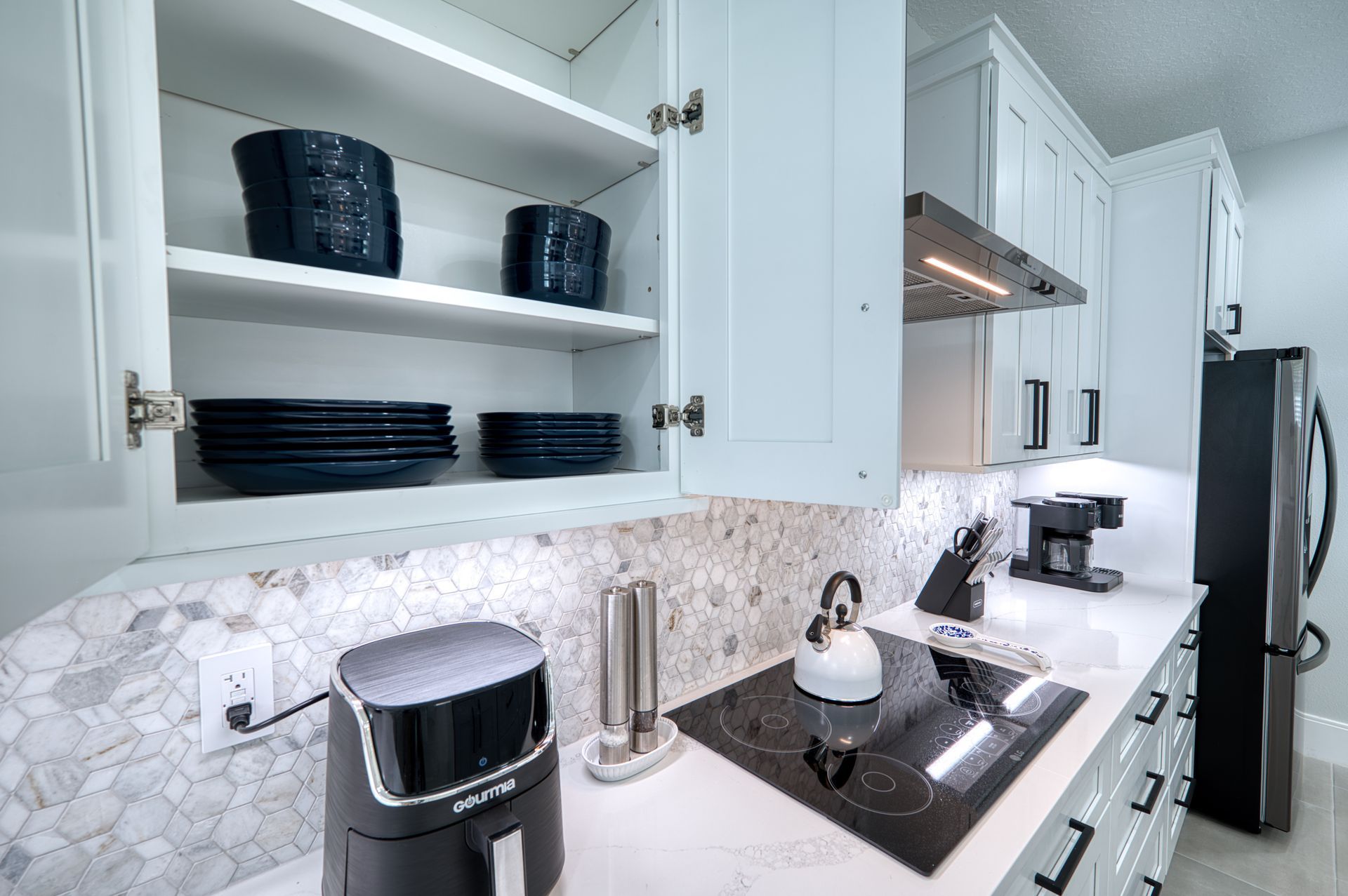White kitchen with open cabinets, black dishes, marble backsplash, and coffee maker on a countertop.
