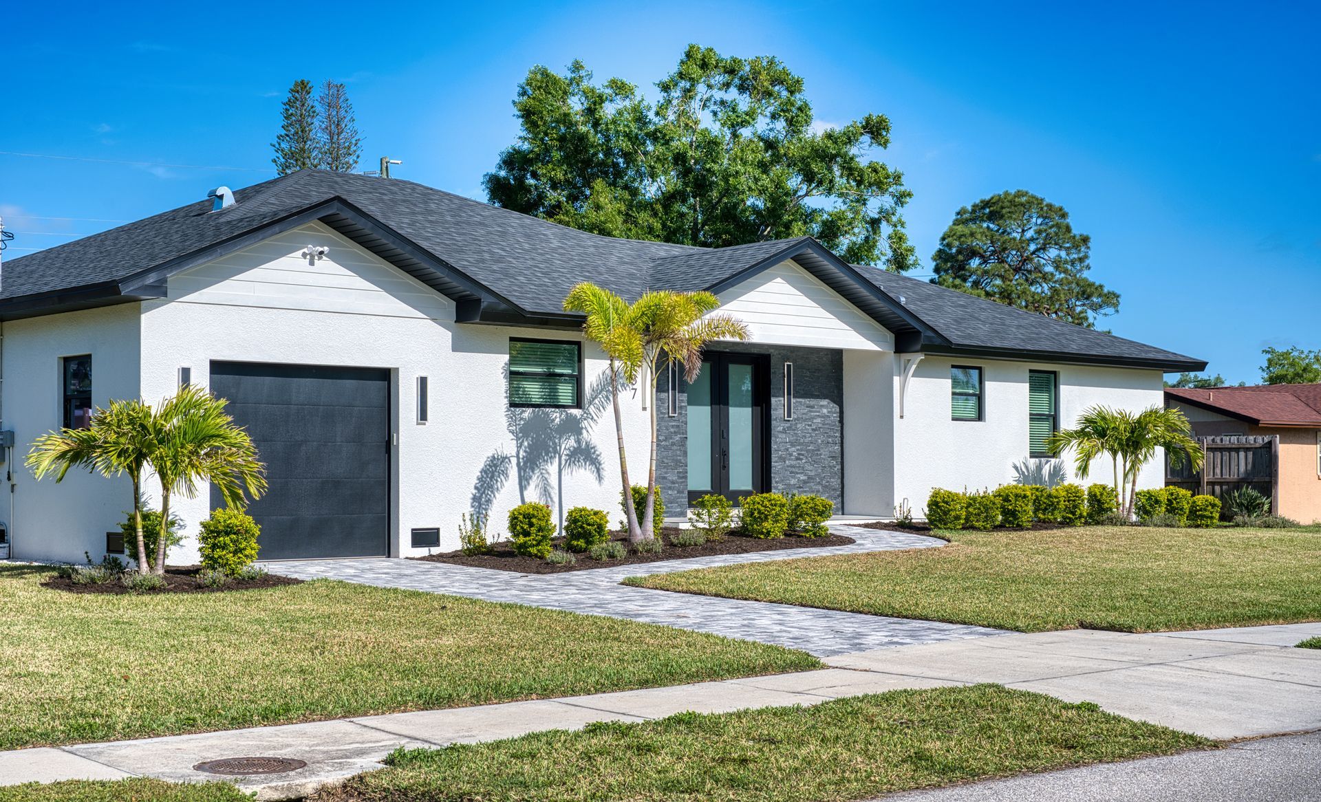 Modern white single-story house with dark gray roof, attached garage, and front lawn on a sunny day