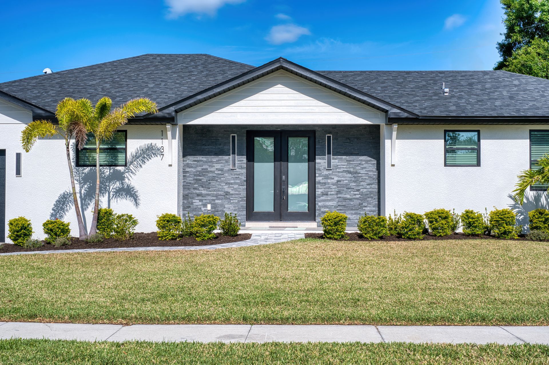 Modern single-story white house with dark gray roof, front lawn, and palm trees under a blue sky