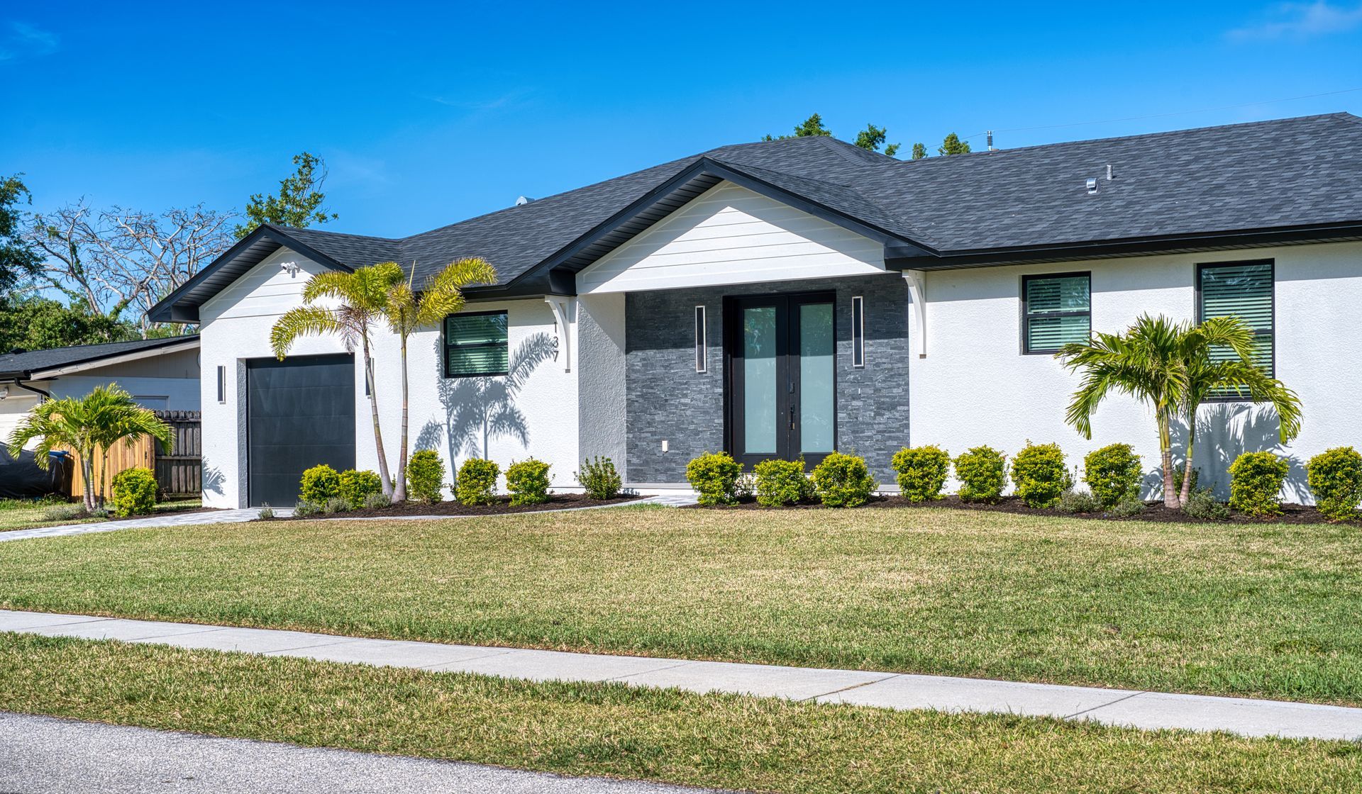 Single-story white suburban house with dark roof, trimmed lawn, and palm landscaping