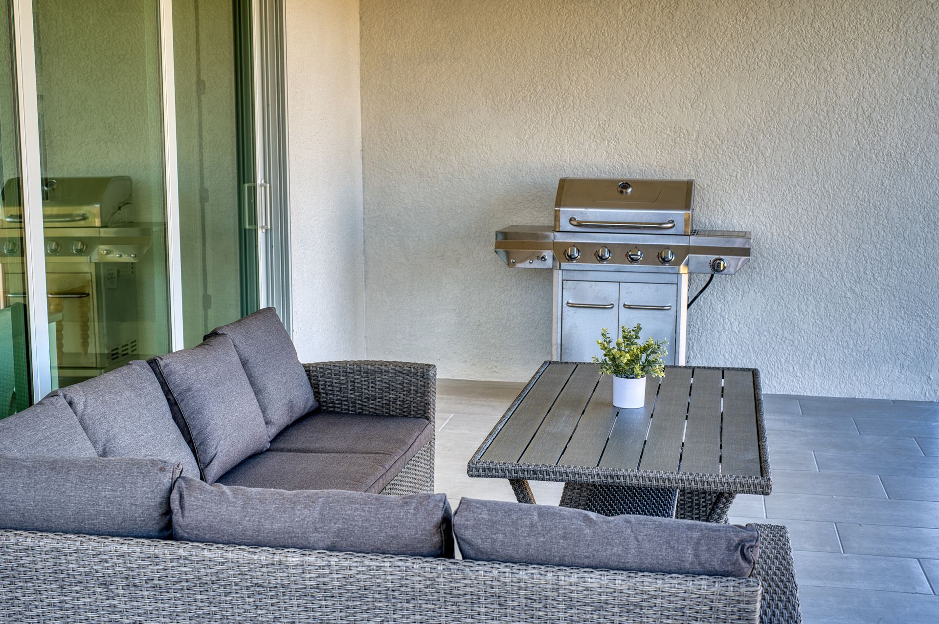 Patio with gray wicker sofa, wooden coffee table, and stainless steel grill against a stucco wall
