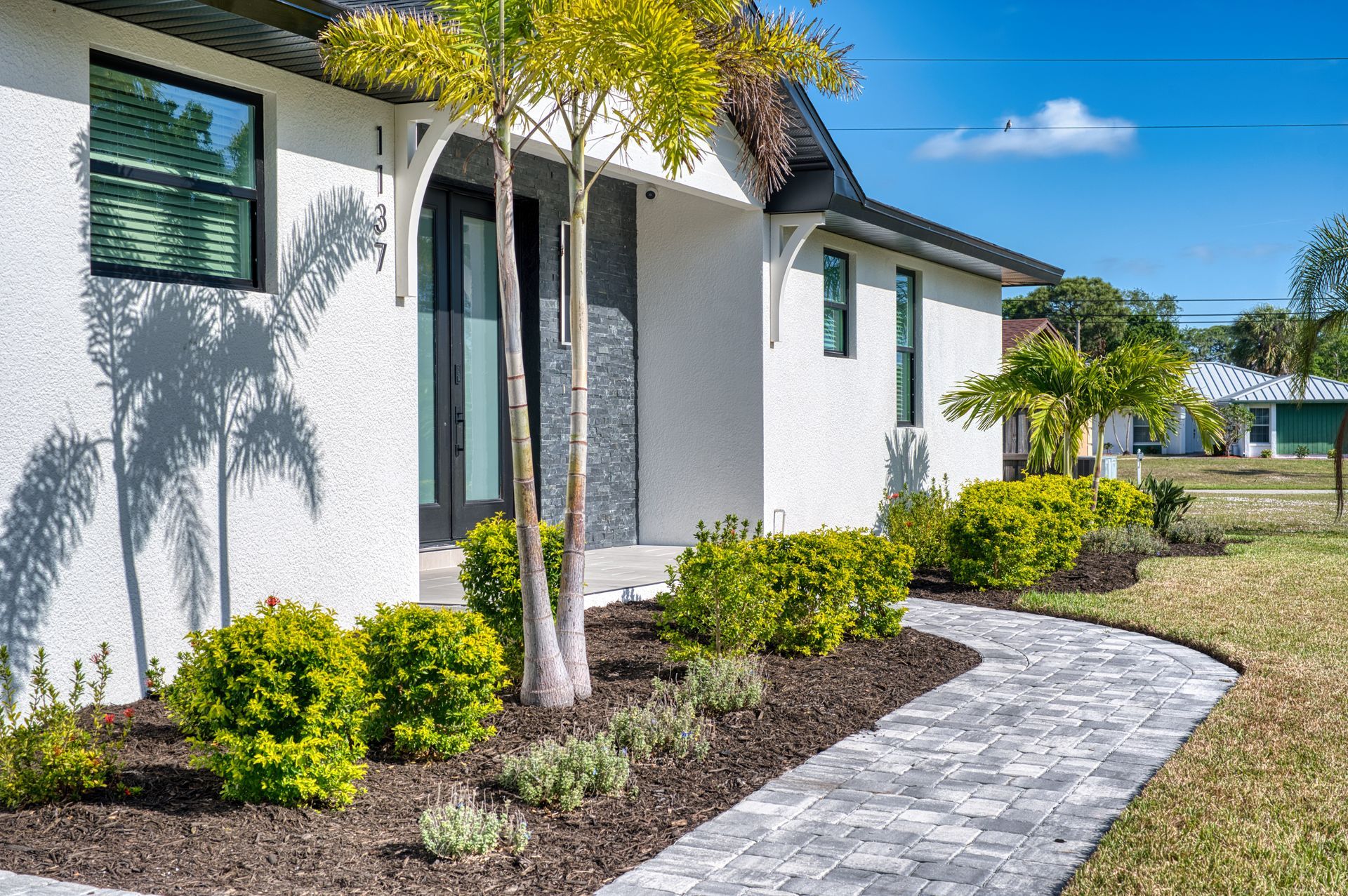 White house exterior with curved stone walkway, trimmed shrubs, and young trees in a sunny yard