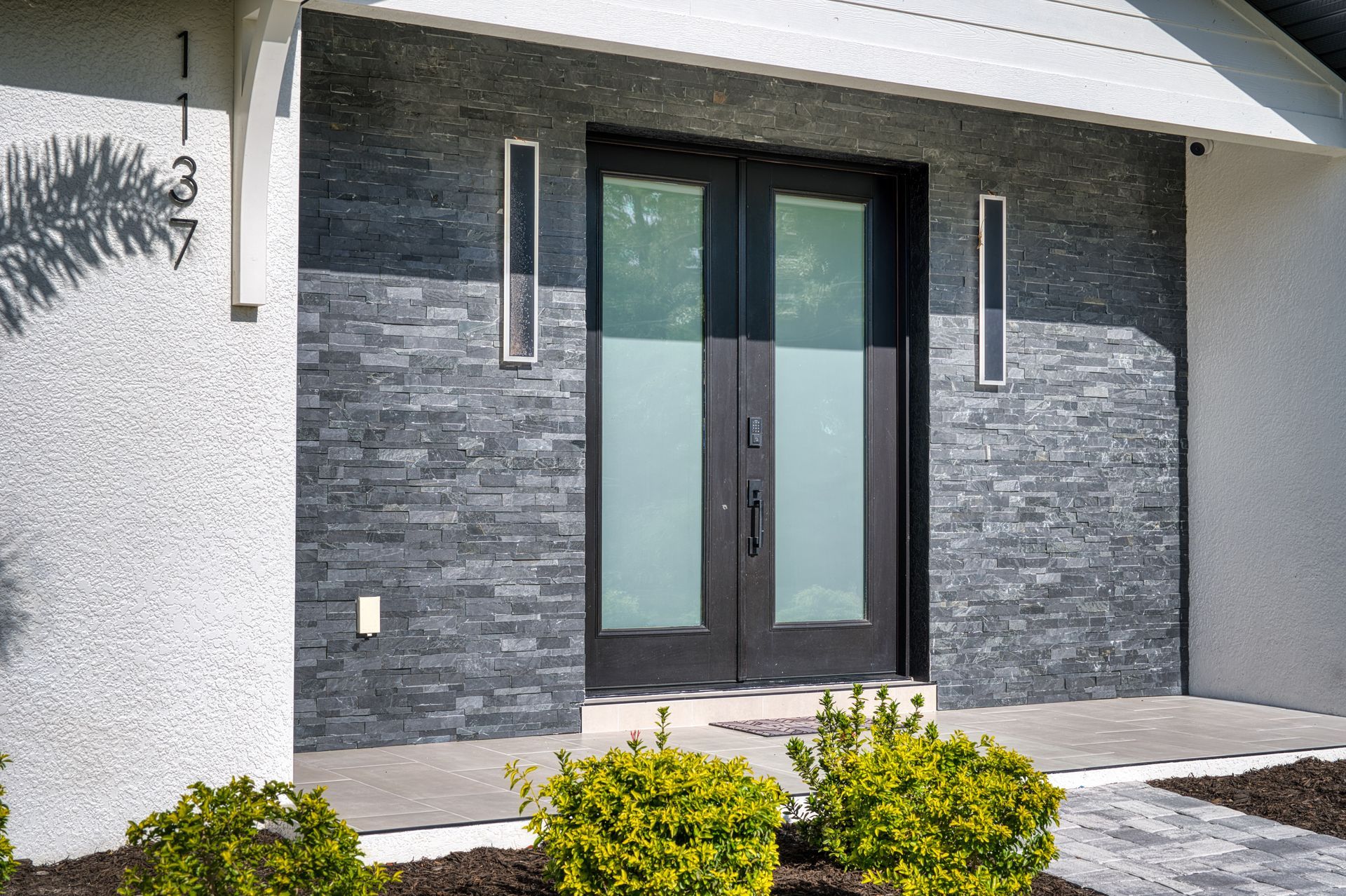 Modern house entrance with dark stone facade, glass double doors, and trimmed shrubs in front