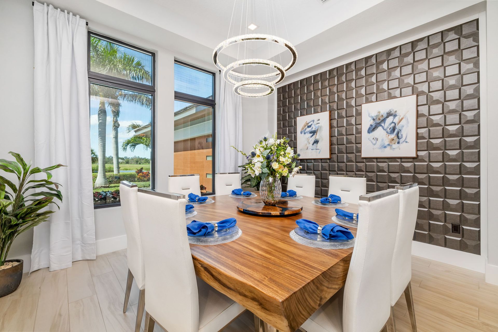 Modern dining room with white chairs, wooden table, blue accents, and a view of palm trees outside.