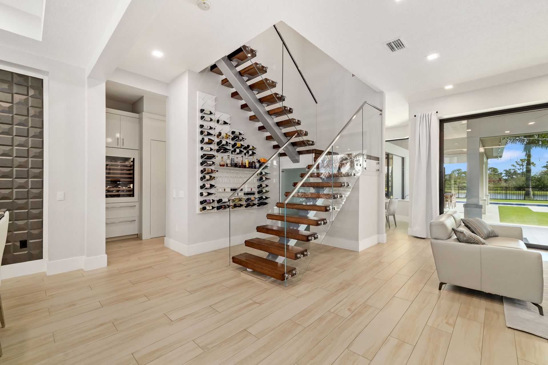 Modern open-concept living room with floating staircase, light wood floors, and large windows overlooking a yard