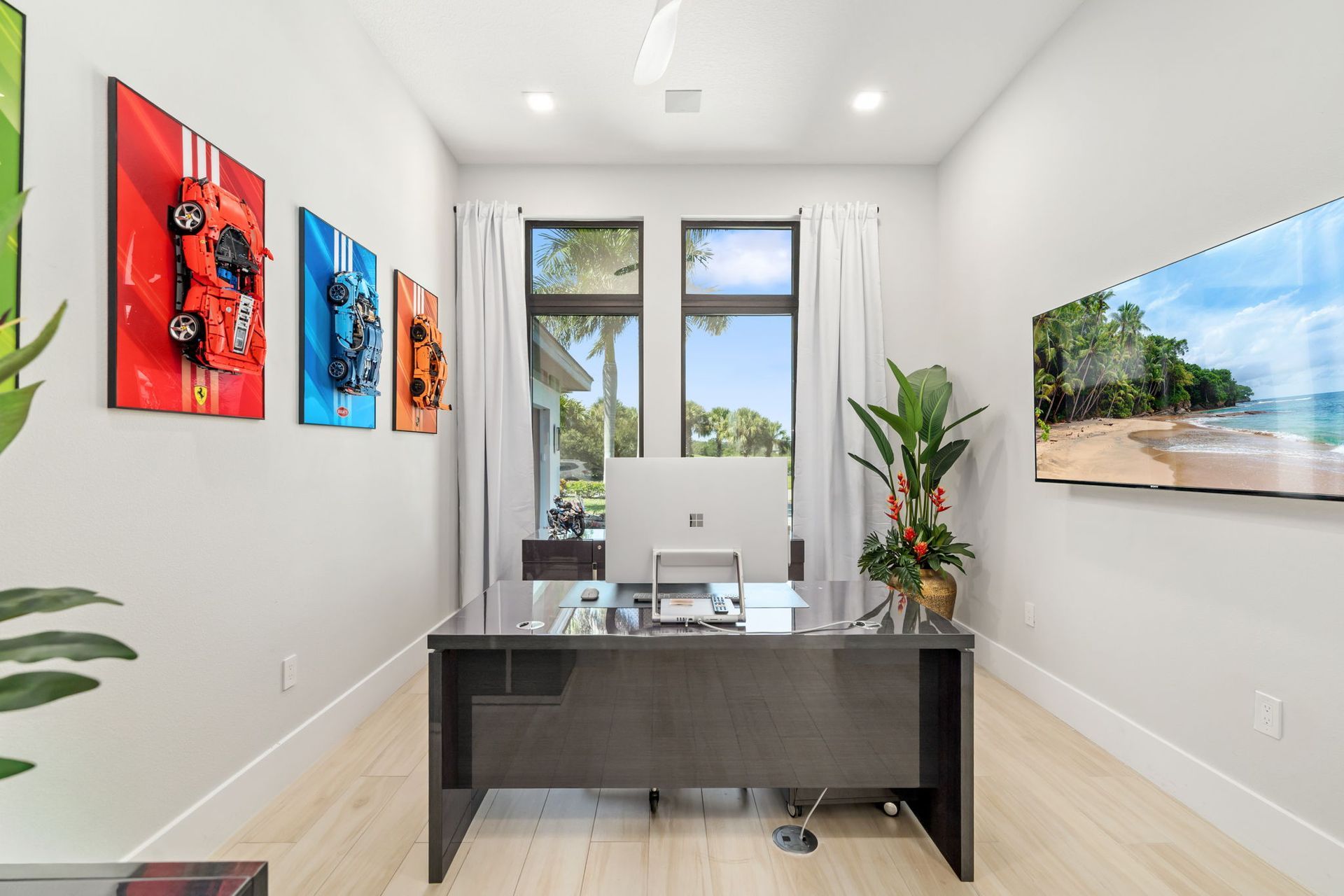 Bright modern office with a desk, dual monitors, wall art, plants, and large windows overlooking palm trees.