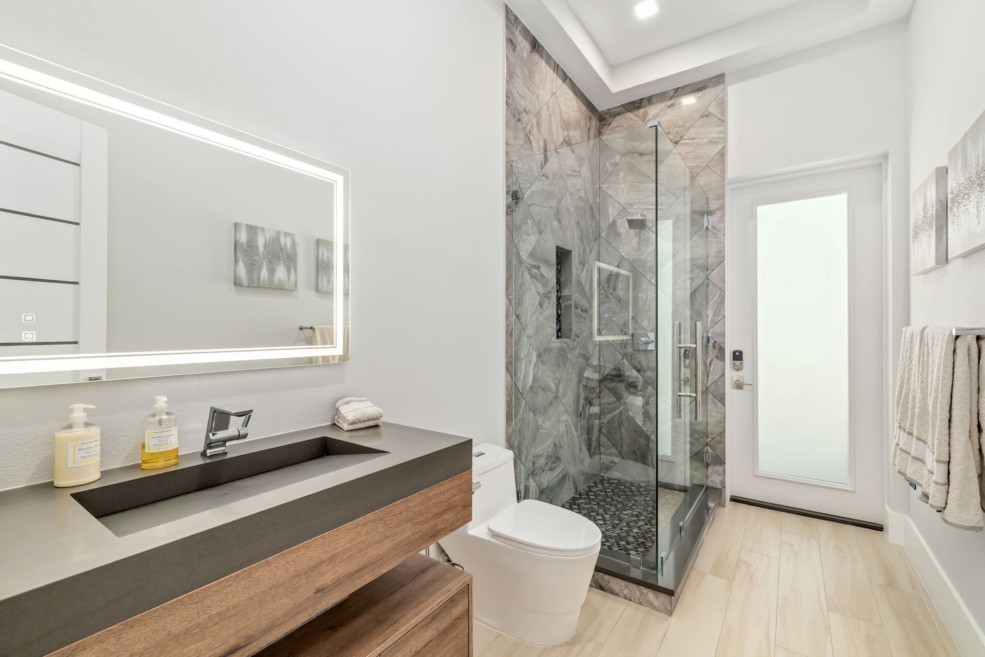 Modern bathroom with double vanity, large mirror, glass shower, and white toilet on light wood flooring