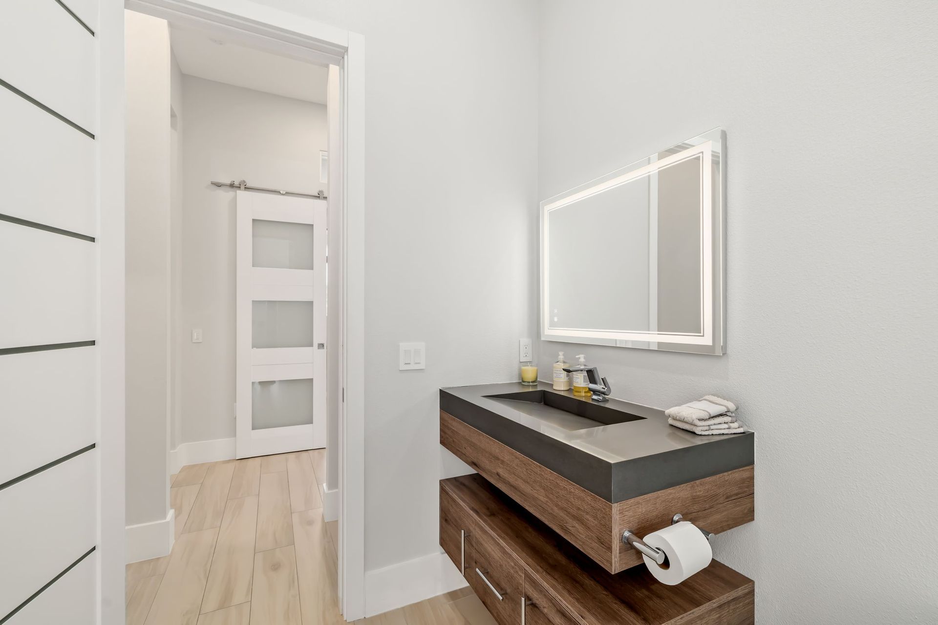 Bright bathroom vanity with dark wood drawers, mirror, and toilet paper holder in a modern white hallway