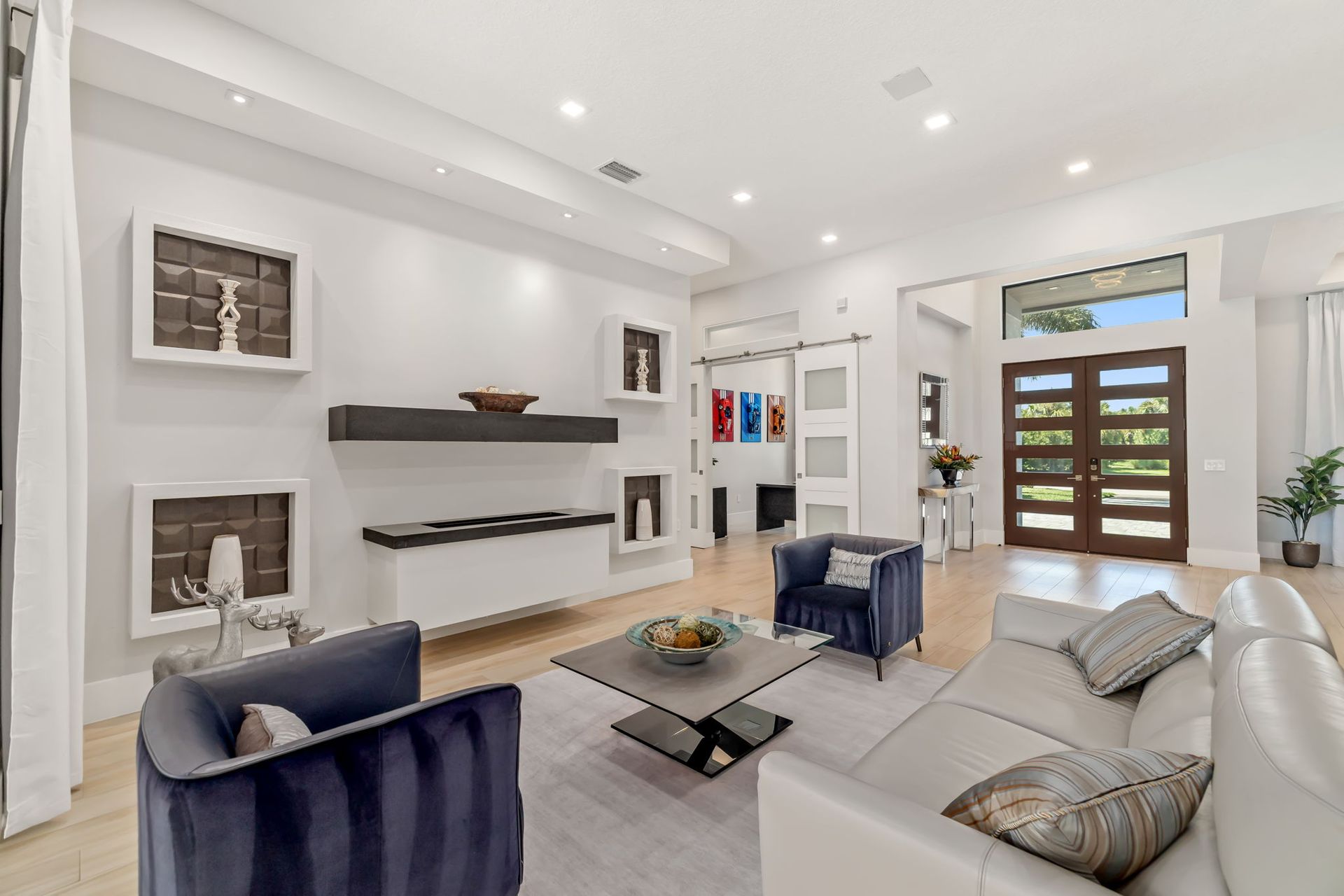 Bright modern living room with white walls, blue chairs, a beige sofa, and large glass front doors.