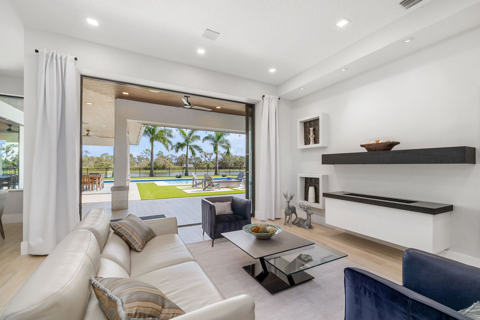 Bright open living room with white sofas, a fireplace, and a view of a pool and palm trees outside.