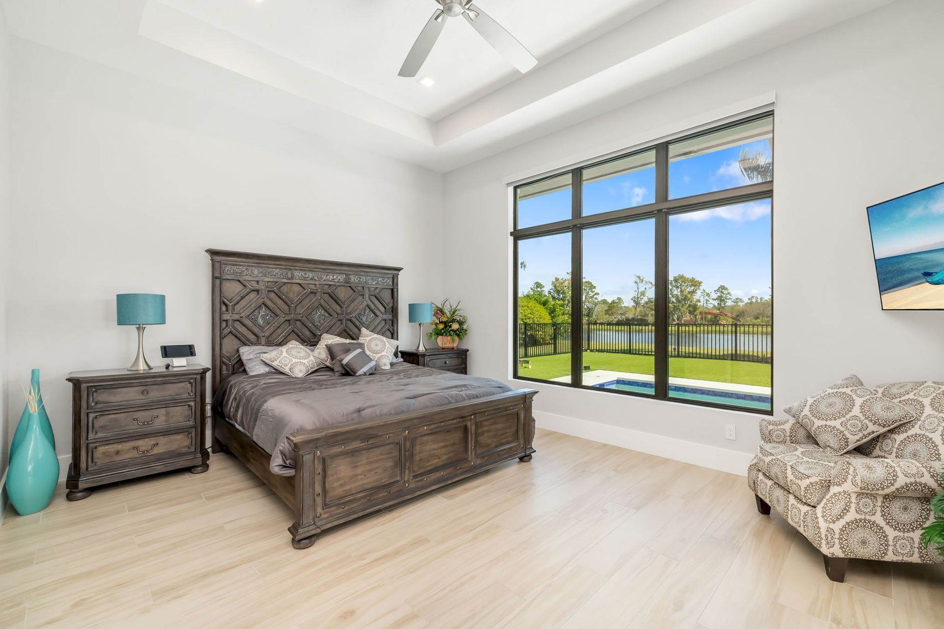 Bright modern bedroom with wood bed, patterned chair, turquoise accents, and large window overlooking a pool.