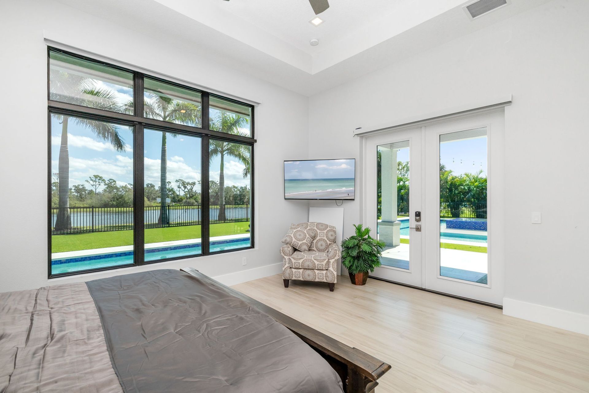 Bright modern bedroom with large windows, white walls, a bed, armchair, TV, and view of a pool outside