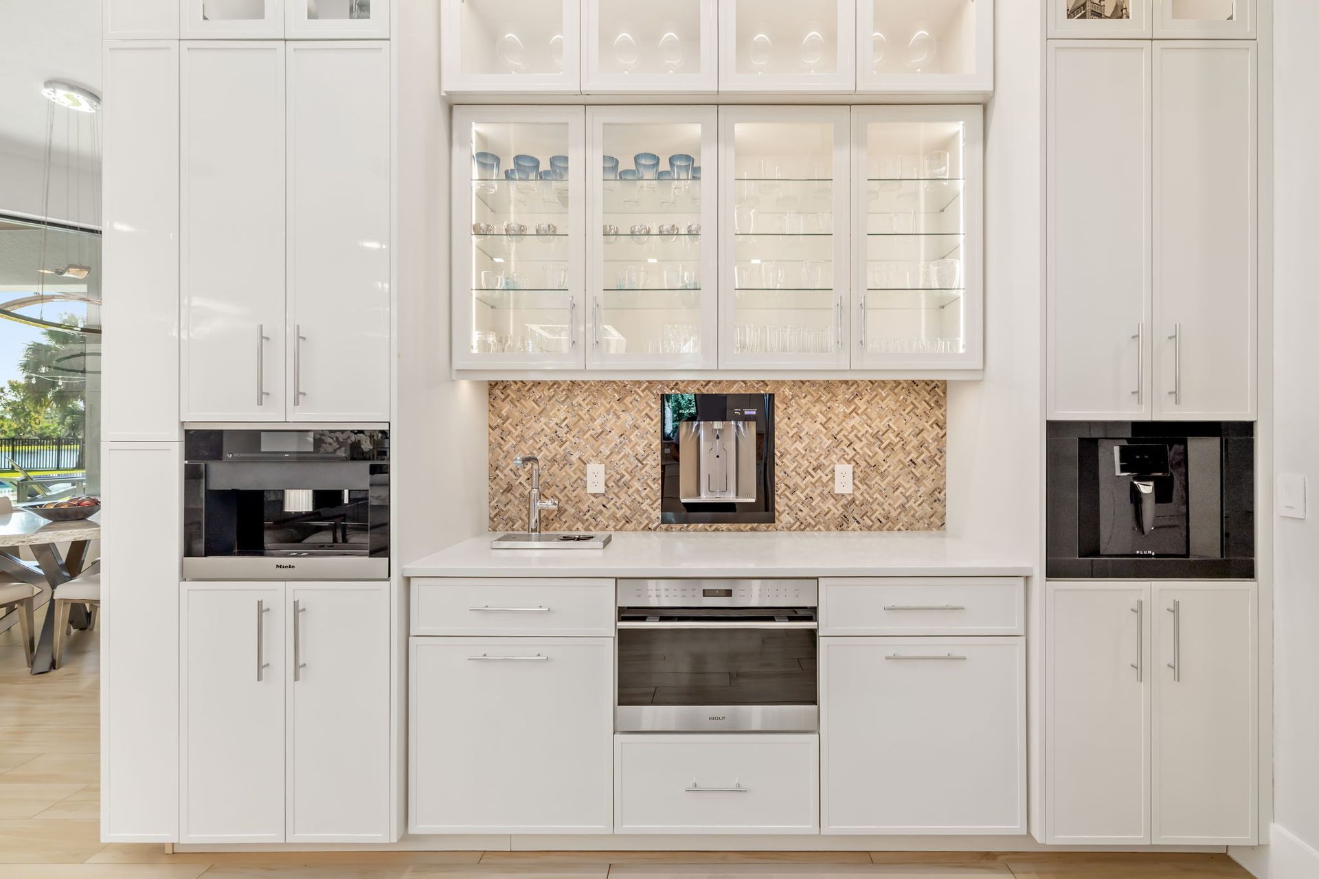 Modern white kitchen with built-in cabinets, glass display doors, coffee machine, and stainless-steel oven.