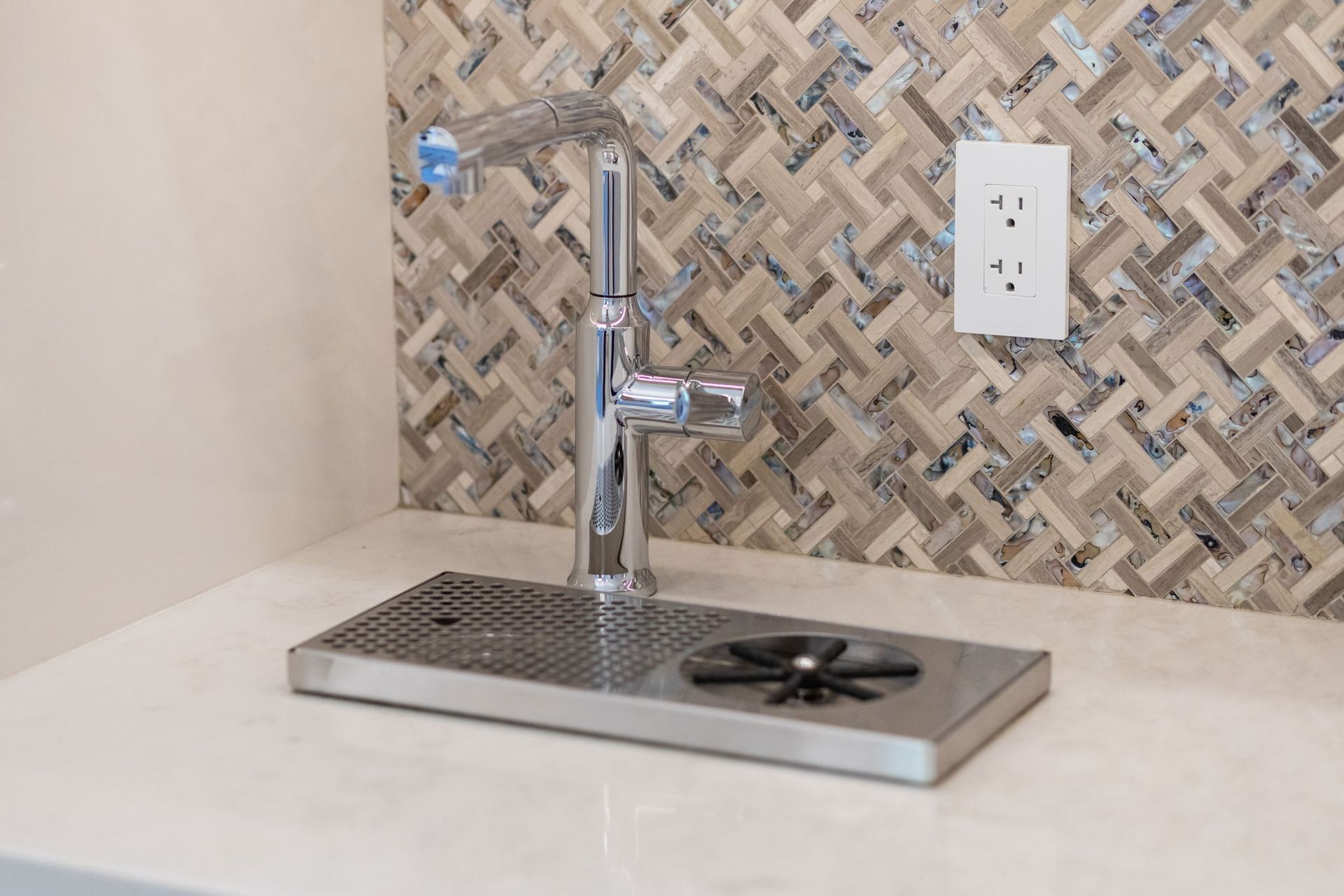 Modern chrome kitchen faucet above a stainless sink on a countertop with a herringbone tile backsplash and outlet