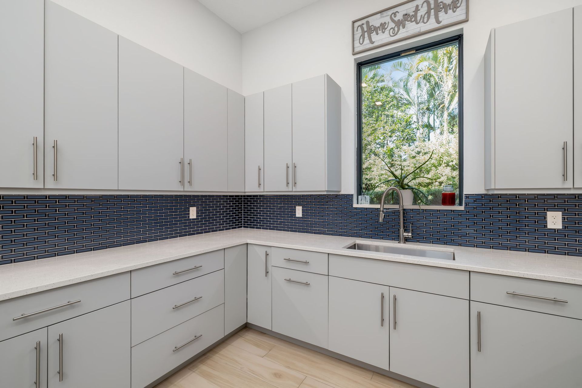 Bright kitchen with white cabinets, blue tile backsplash, and a window above the sink overlooking trees.