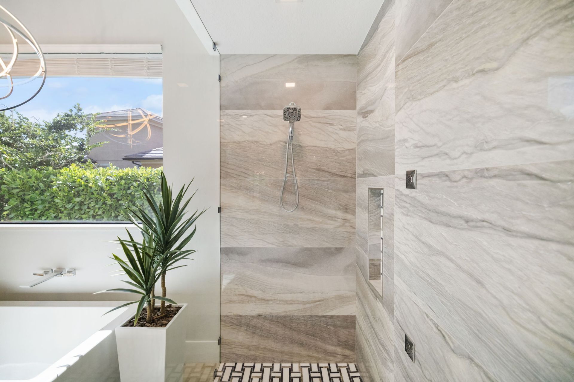Modern shower with beige tile, mosaic floor, and a white bathtub beside a window and potted plant