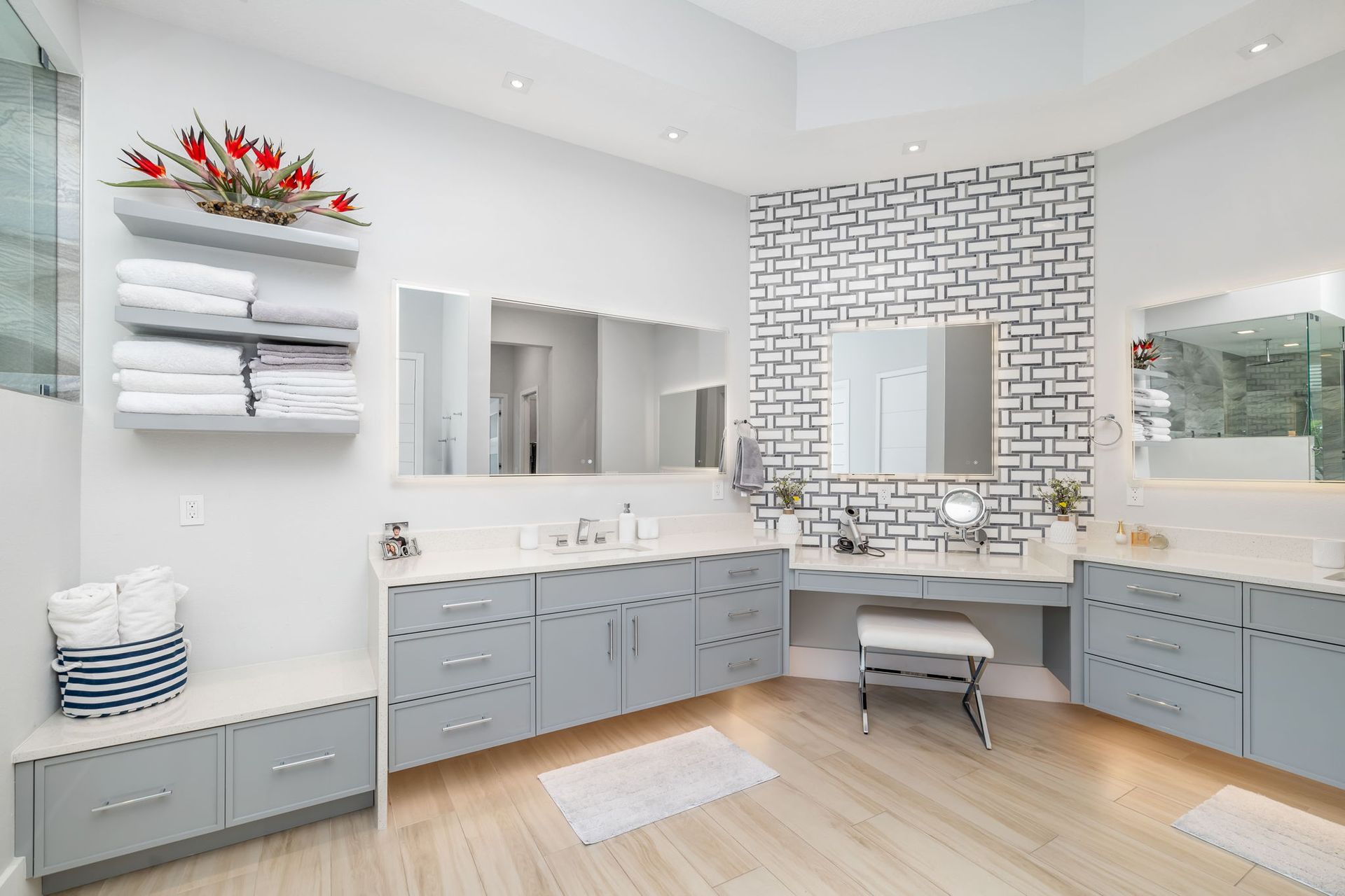 Bright modern bathroom with double sinks, gray cabinets, white tile, and a bench by a large mirror.