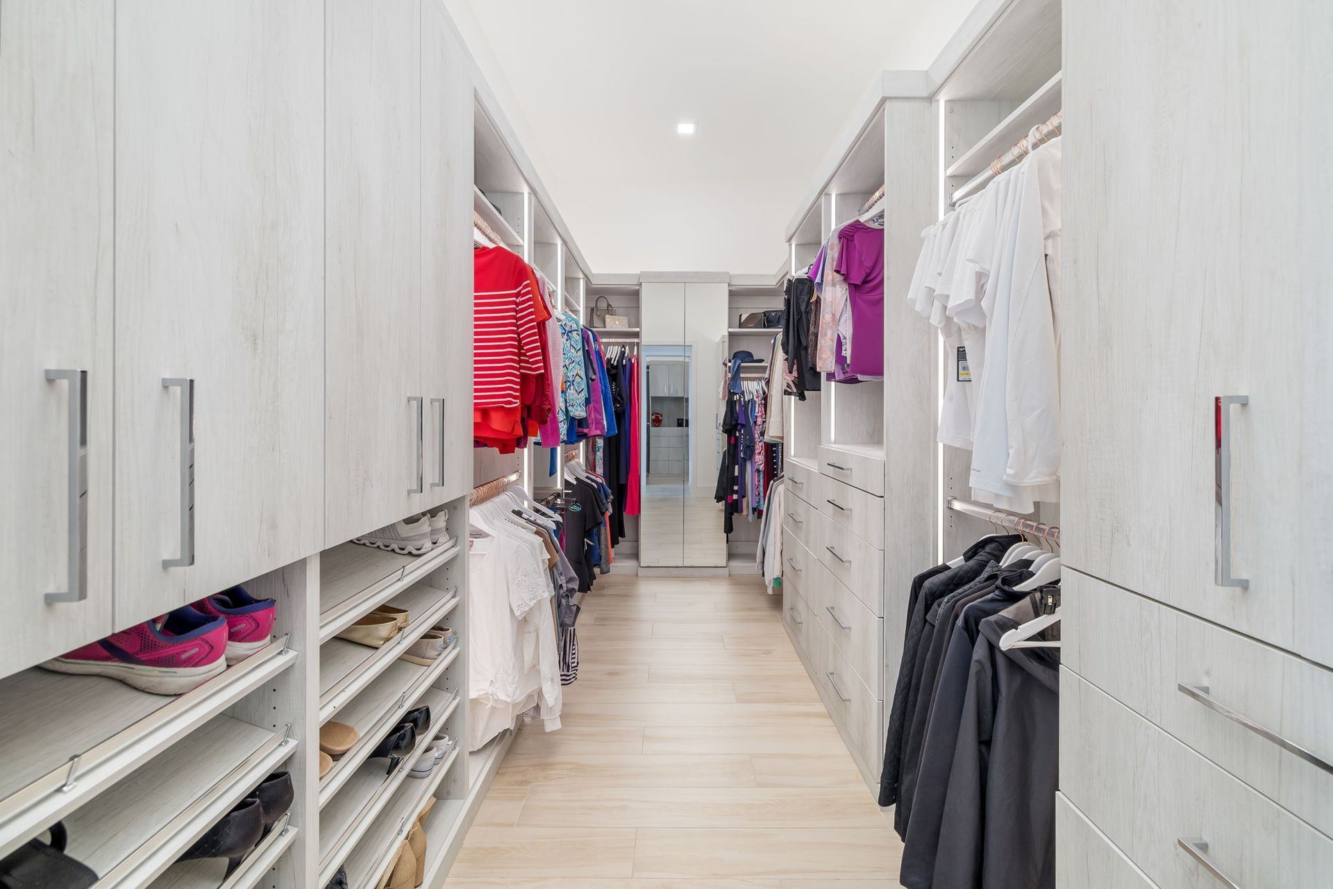 Bright walk-in closet with white cabinets, hanging clothes, shelves, and light wood flooring